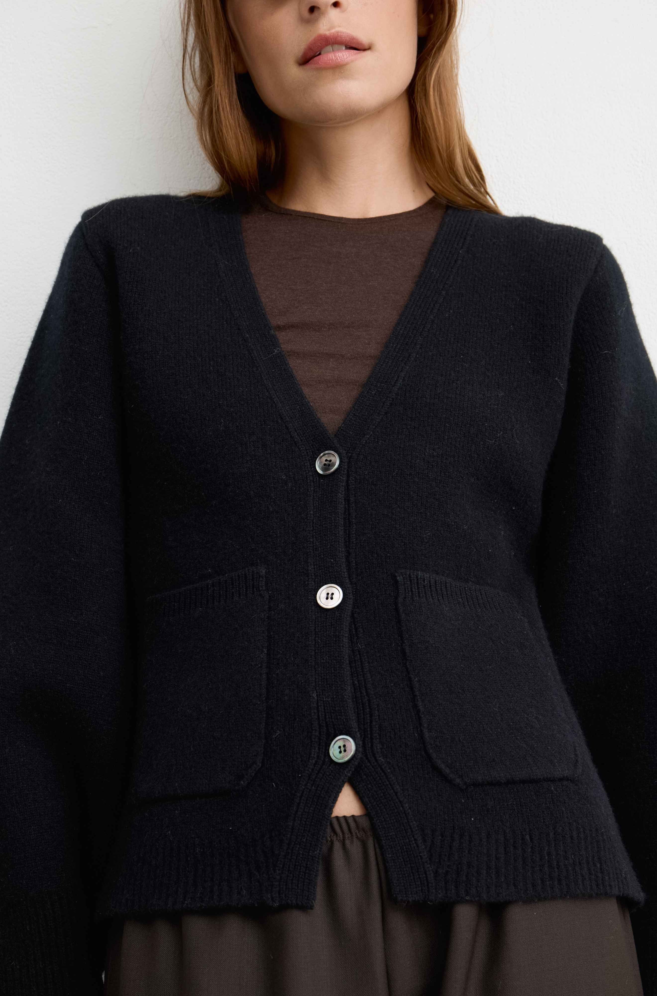 A close-up of a woman wearing a black knit cardigan with three silver buttons over a brown top, shown from the chest up against a plain light background.