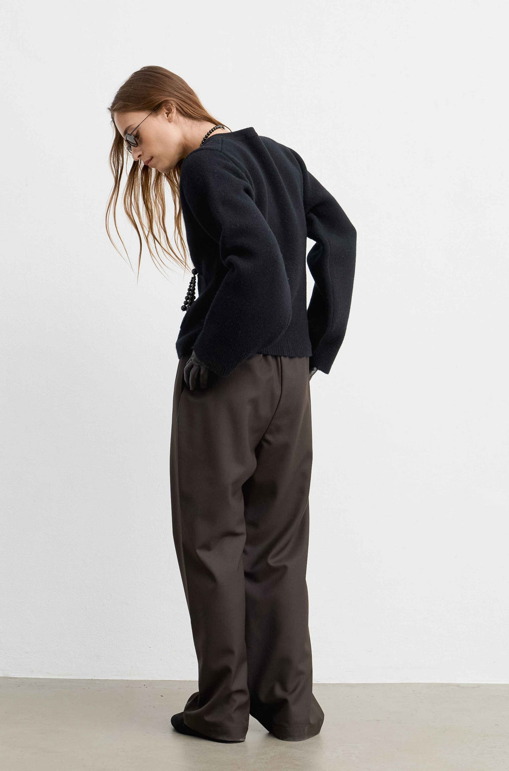 A model wearing a black cropped knit sweater and dark brown wide-leg trousers, photographed from the back against a plain white studio background.