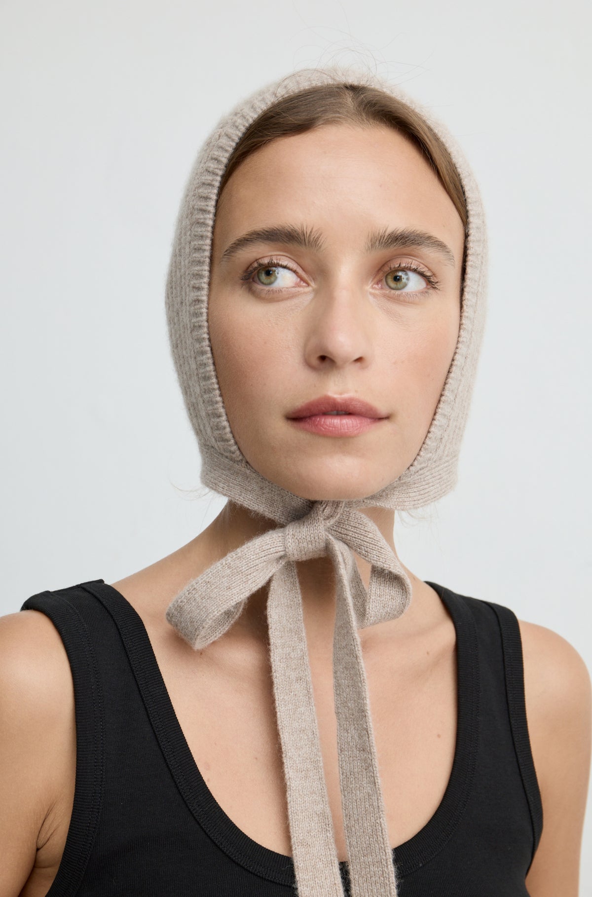 Woman wearing a light brown knit hat tied around the neck with a black tank top against a light grey wall