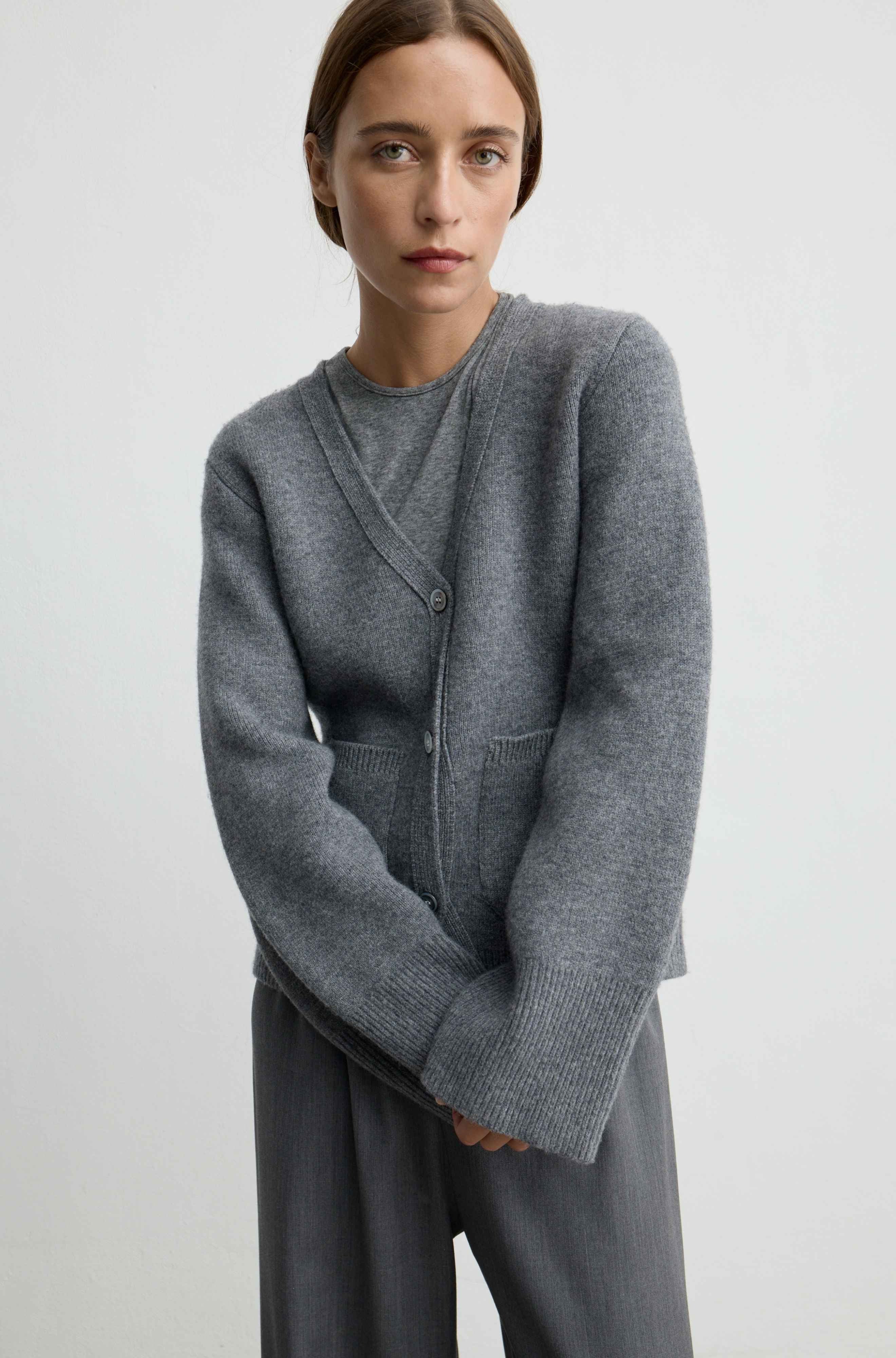 A woman wearing a gray knit cardigan with a split V-neck and front buttons, paired with gray wide-leg trousers, photographed against a plain light gray studio background.