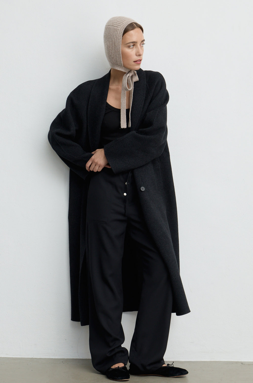 Woman wearing a light brown knit hat tied around the neck paired with a black coat, top, and pants against a light grey wall