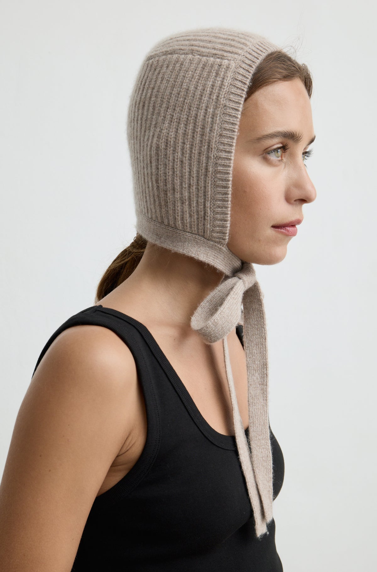 Woman wearing a light brown knit hat tied around the neck with a black tank top against a light grey wall