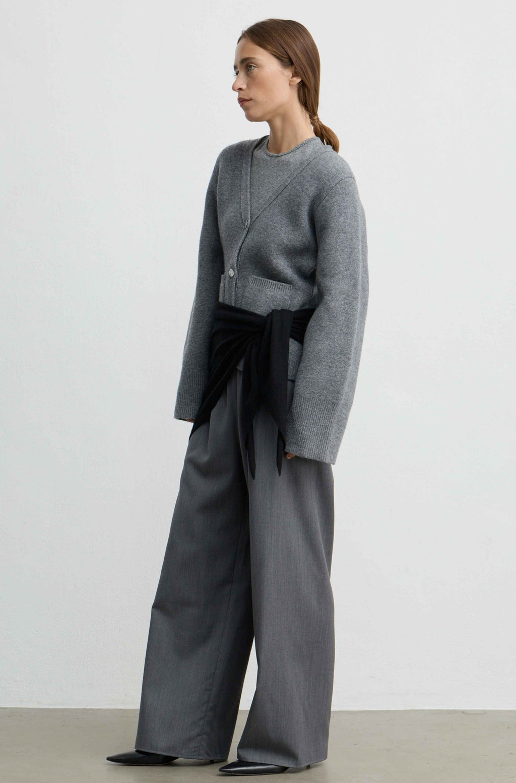 A woman stands in profile wearing a gray knit cropped sweater with a black waist tie and wide-leg gray trousers against a plain light gray studio background.