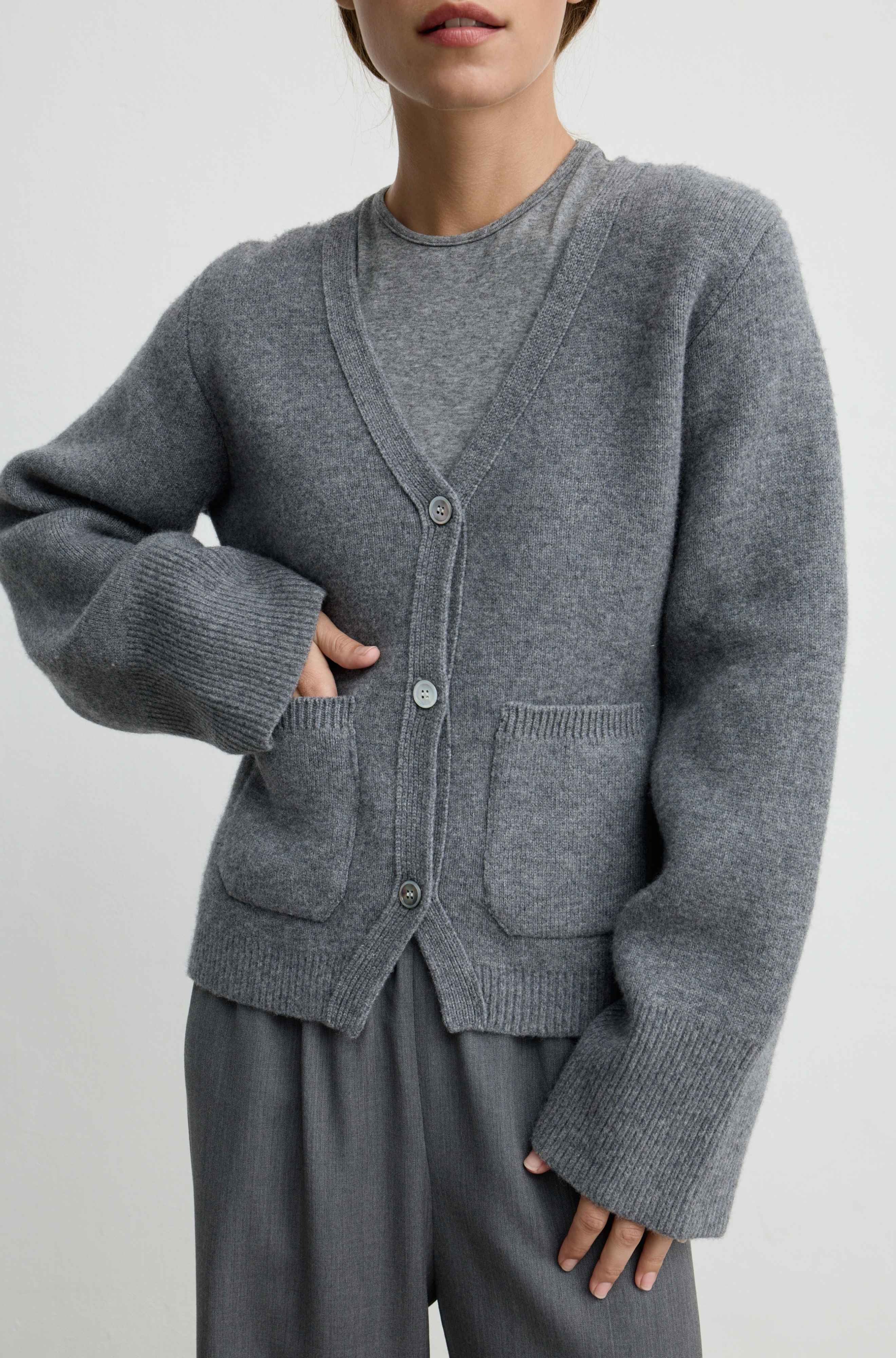 A person wearing a heather gray knit cardigan with a V-neck, front buttons, and large front patch pockets, paired with gray pants against a plain light gray studio background.