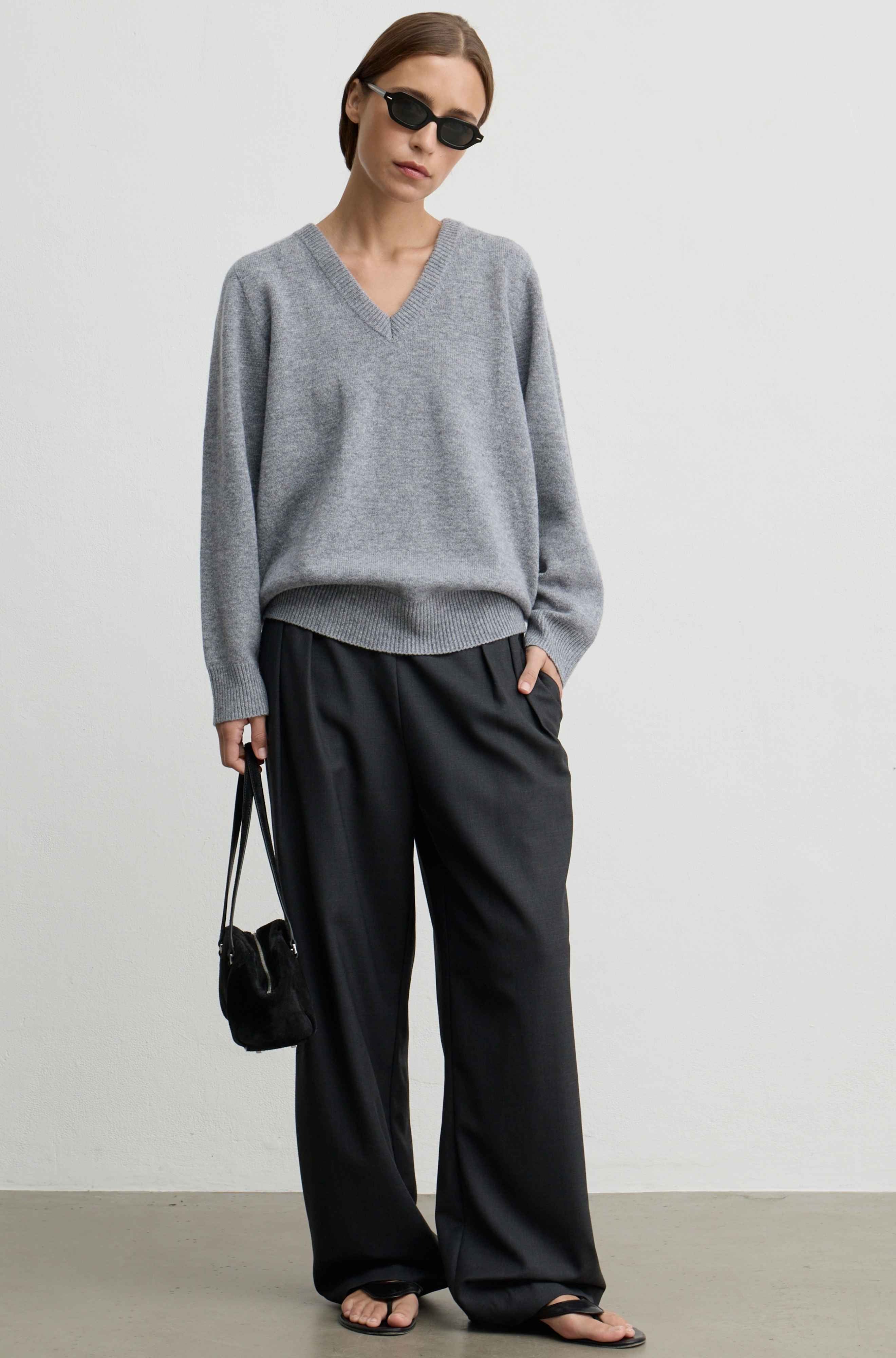 A person wearing loose grey wide-leg trousers with a drawstring waist, paired with a gray sweater and black sandals, holding a black handbag against a plain light gray wall.