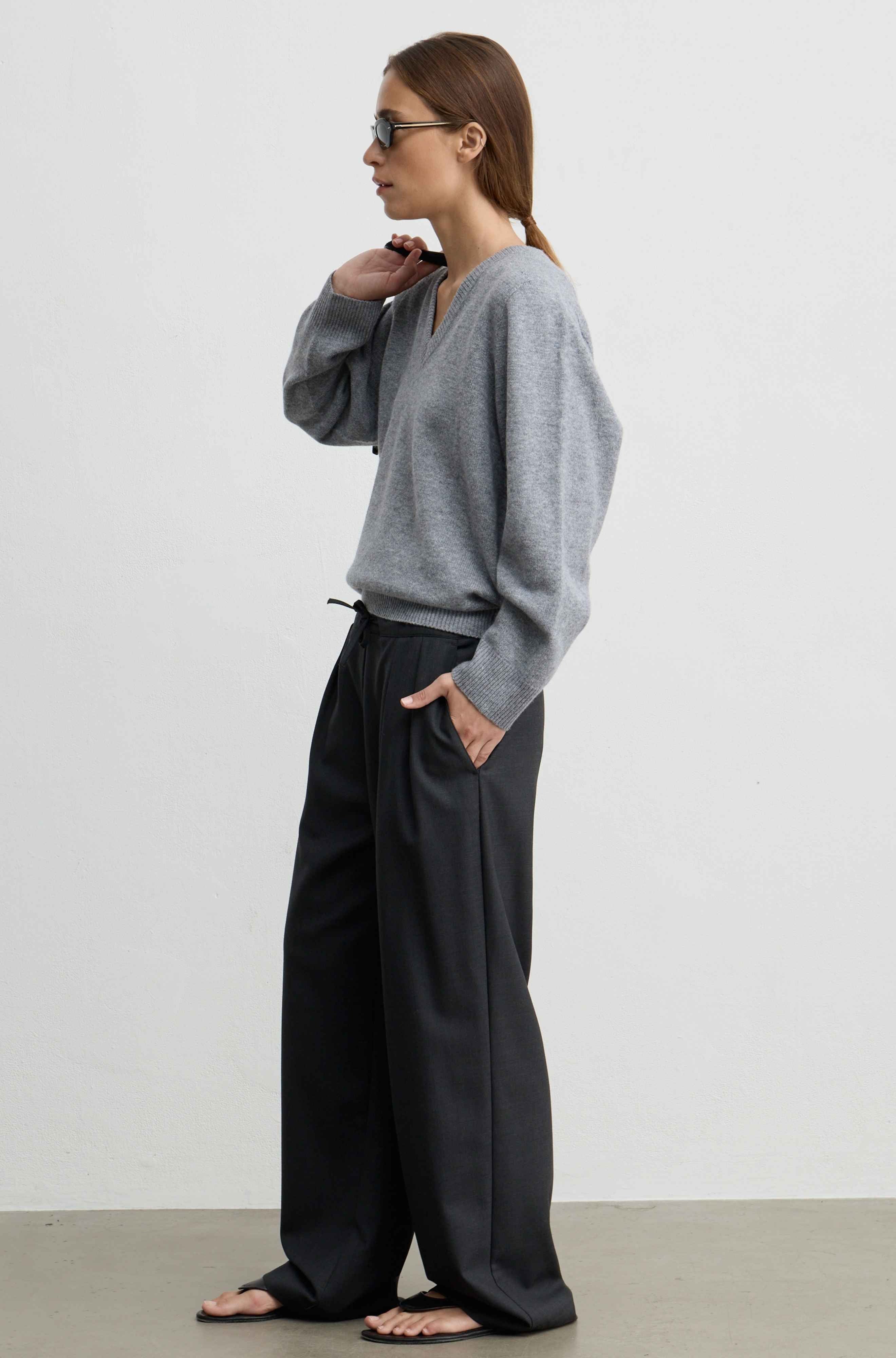 A woman stands in profile wearing a light gray knit sweater and wide-leg grey trousers, with black sandals, against a plain light gray studio background.