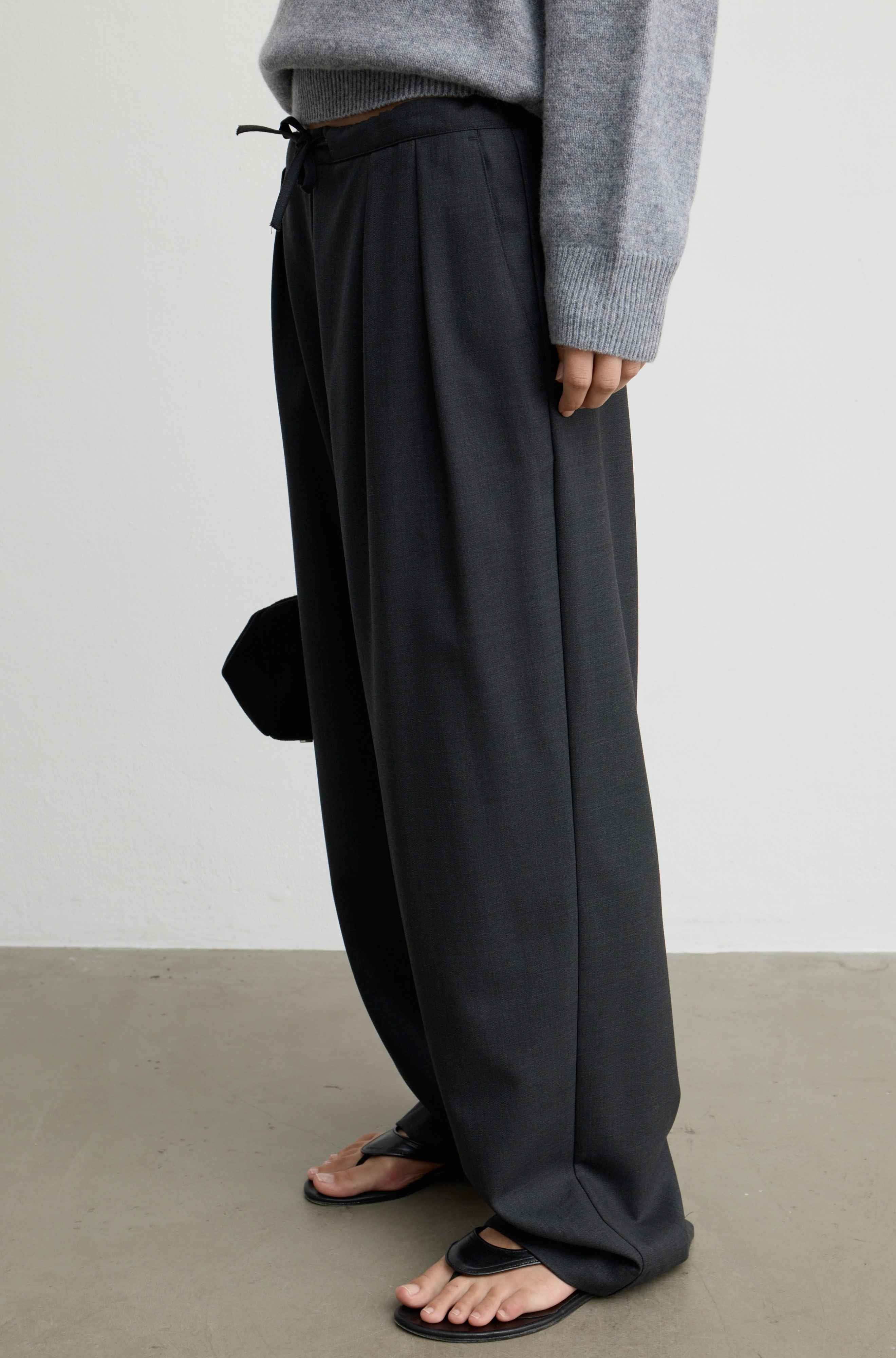 A person wearing loose, wide-leg grey pants with a drawstring waist, styled with a gray knit sweater and black sandals against a plain light gray wall and concrete floor.