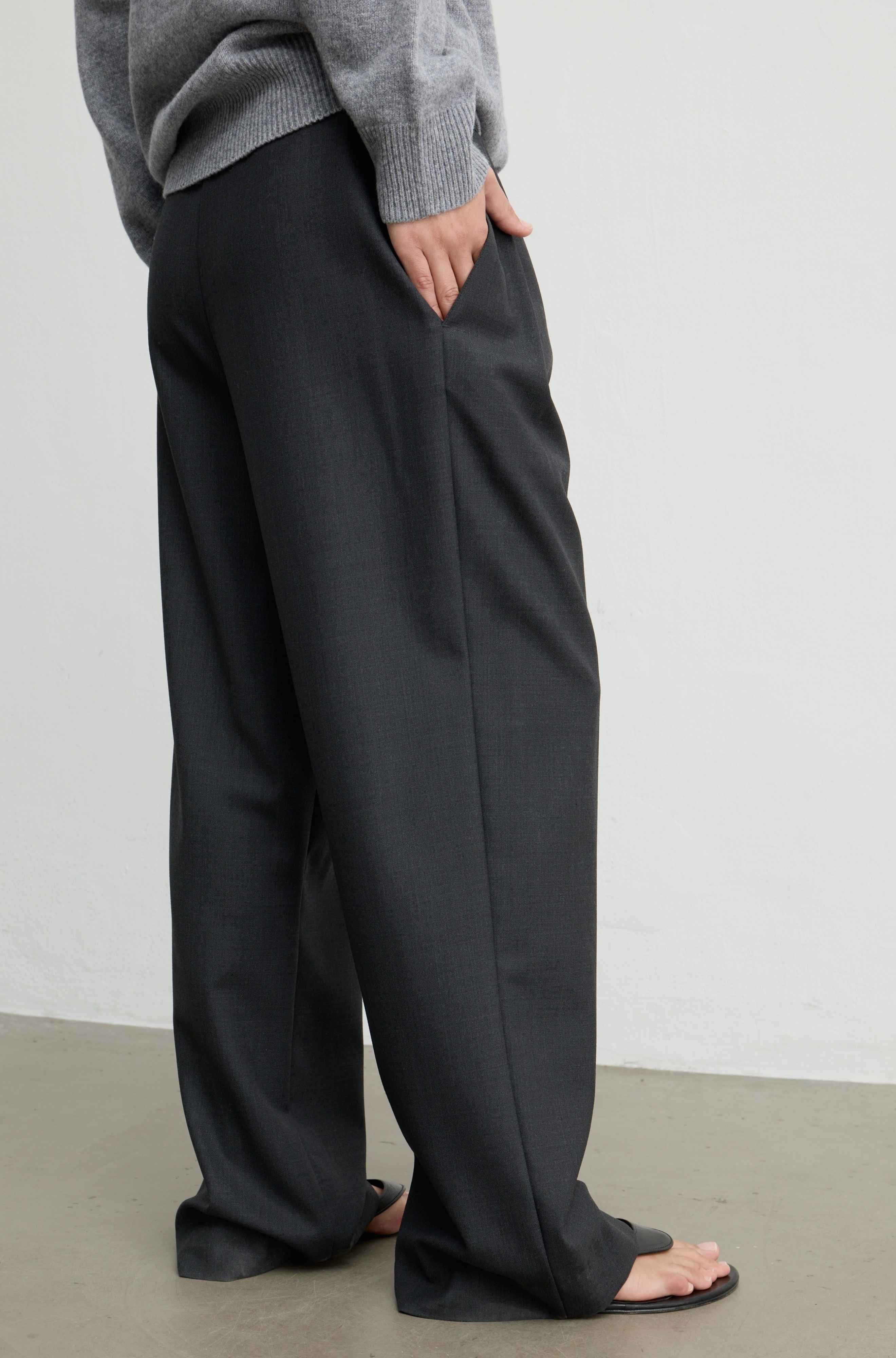 A person wearing loose, wide-leg dark charcoal gray trousers with a gray knit top and black sandals, standing against a plain light gray wall.