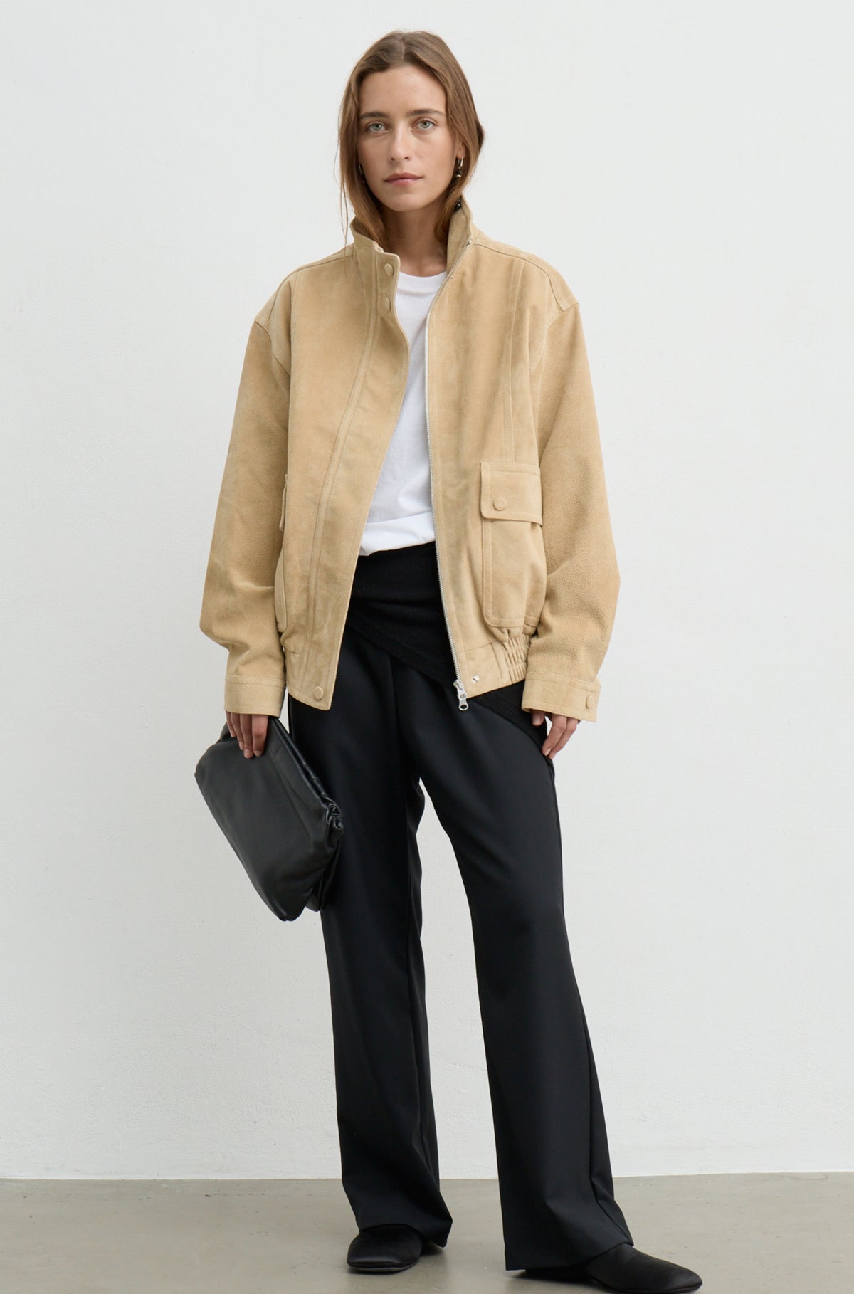A woman stands against a plain light gray studio background wearing a beige suede jacket over a white top and black wide-leg trousers, while holding a black clutch.