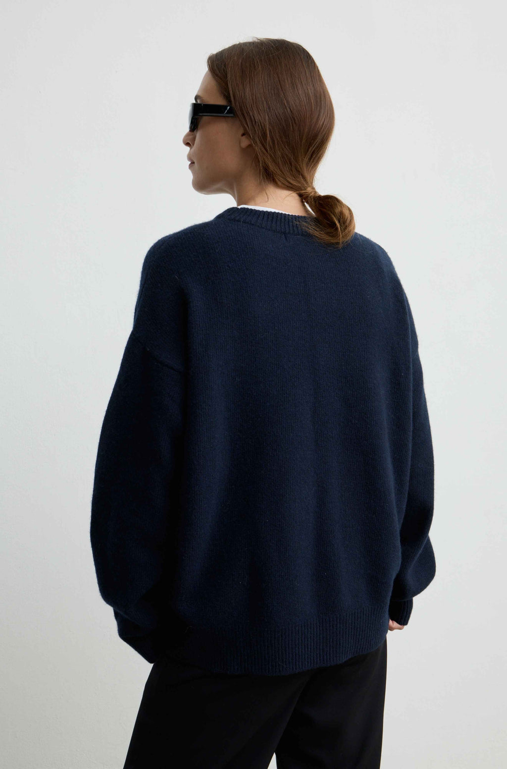 A woman shown from the back wearing an oversized dark navy knit sweater and black pants against a plain light gray studio background.