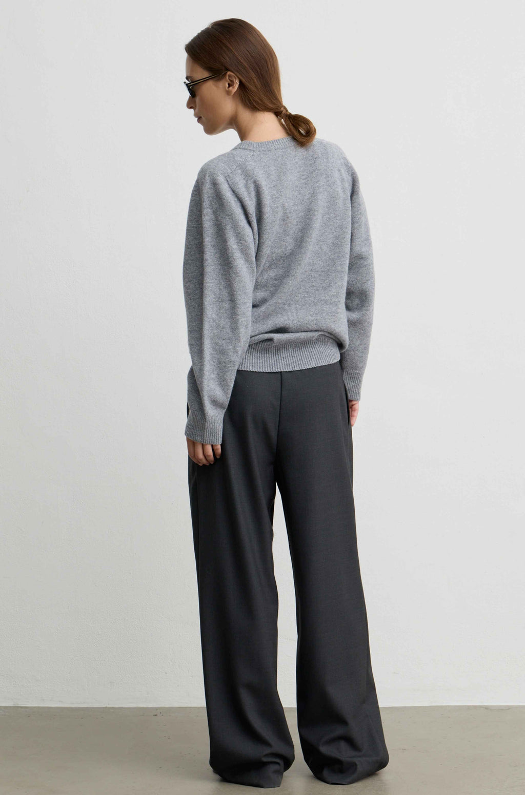 A woman shown from the back wearing a light gray knit crewneck sweater and dark charcoal wide-leg trousers, standing against a plain light gray wall.