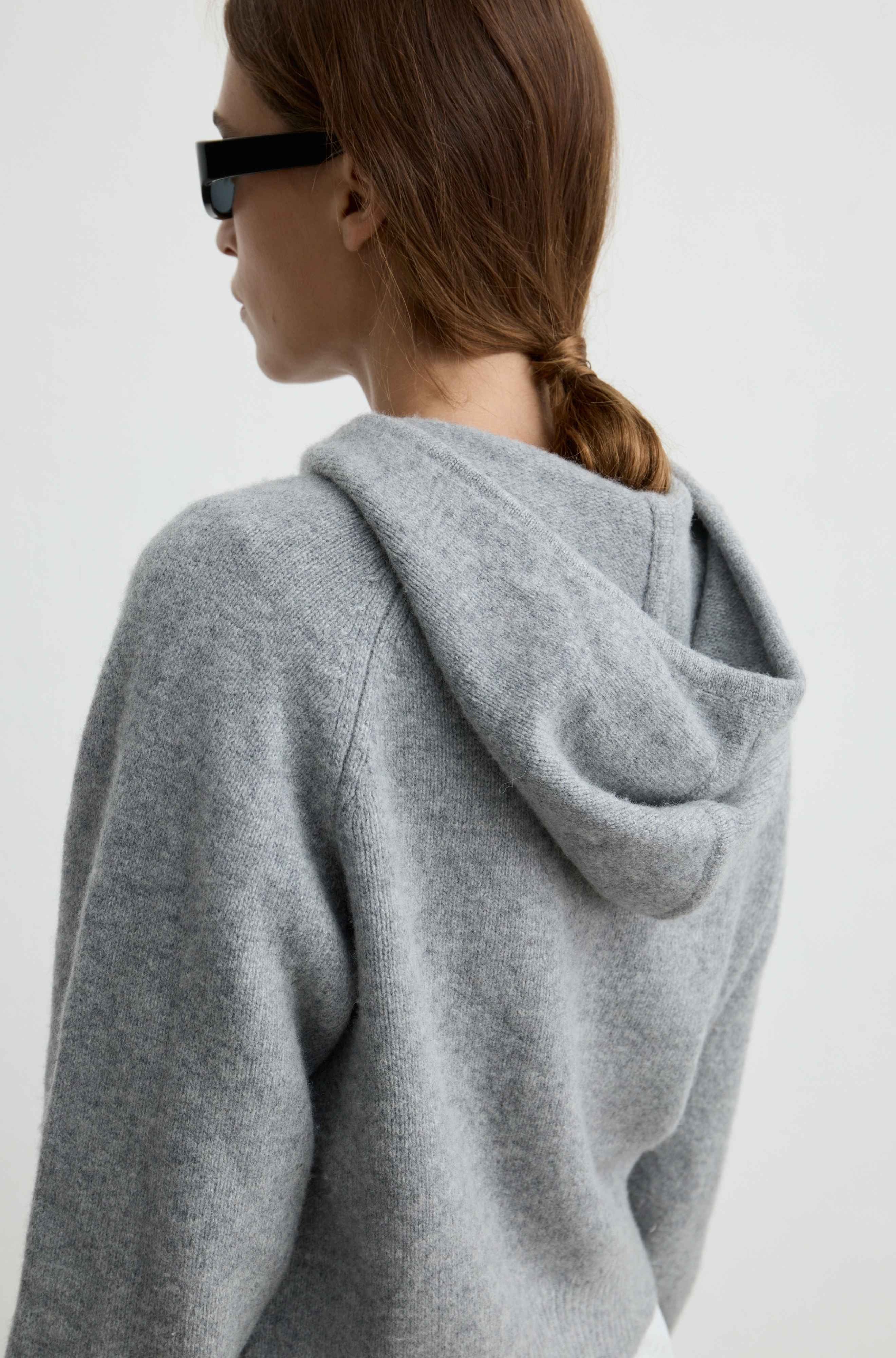 A woman shown from the back wearing a light gray heathered hooded sweatshirt and black sunglasses against a plain white background.