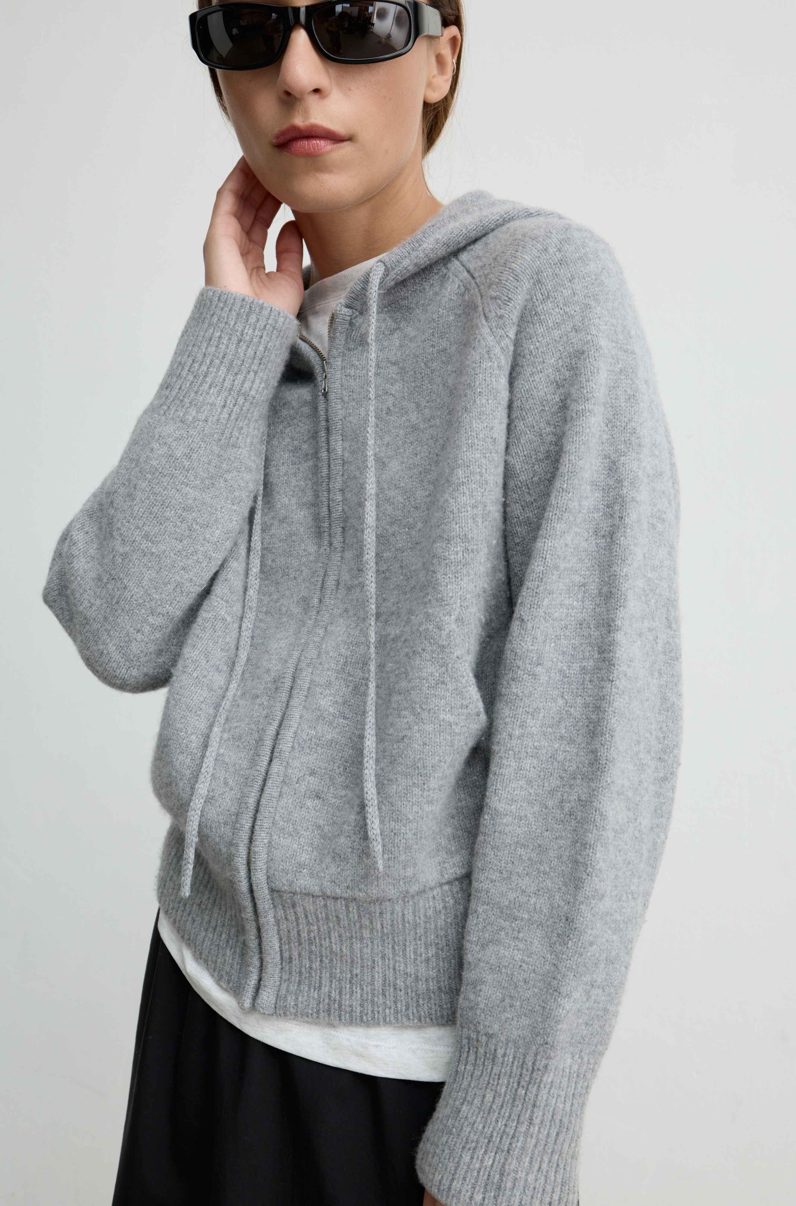 A model wearing a light gray heathered knit zip-up hoodie with drawstrings, styled with black sunglasses and a white undershirt.