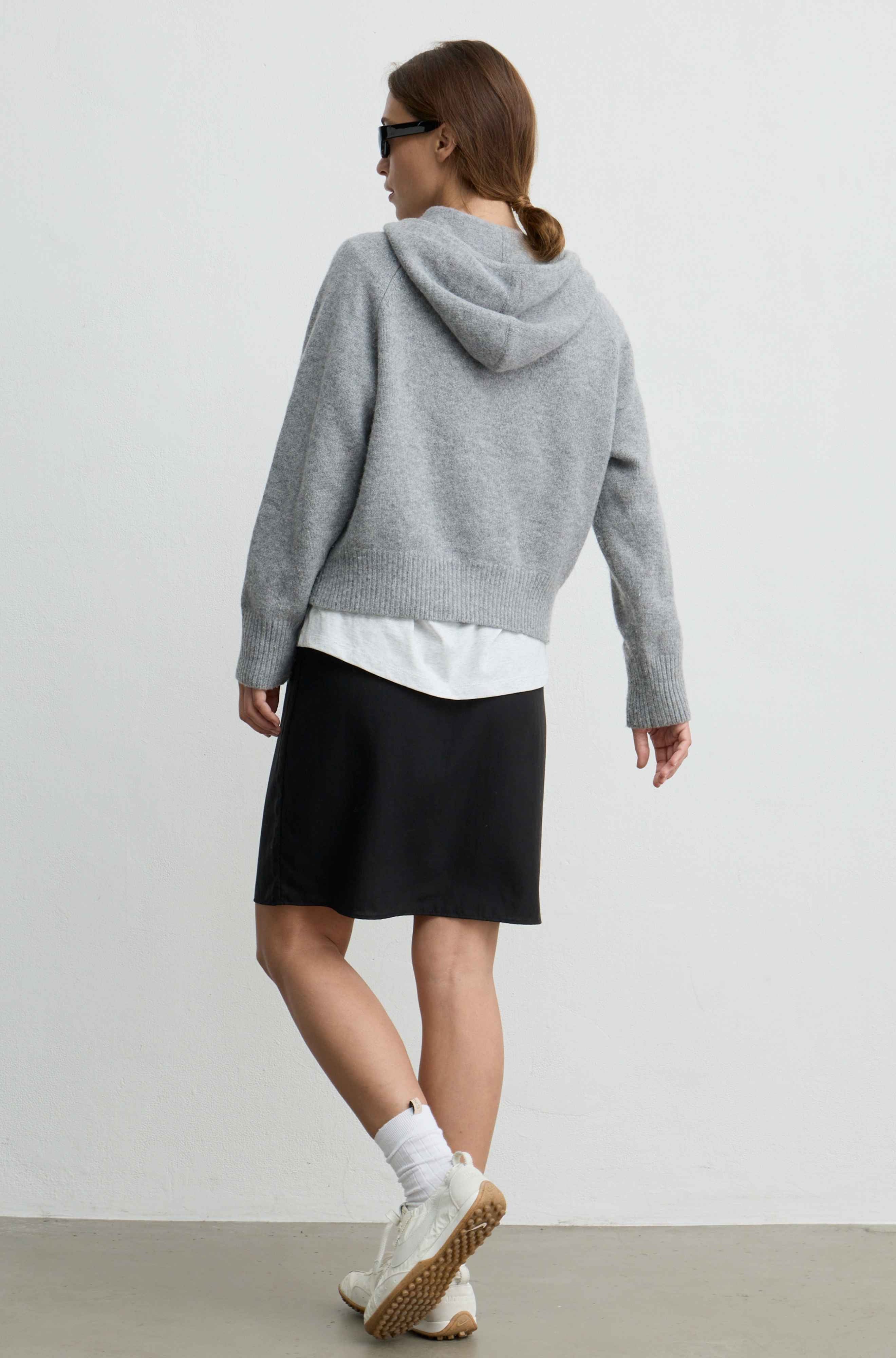 A woman is shown from behind wearing a light gray oversized knit hoodie, a white undershirt, a black skirt, white socks, and white sneakers with brown soles, set against a plain light gray studio background.