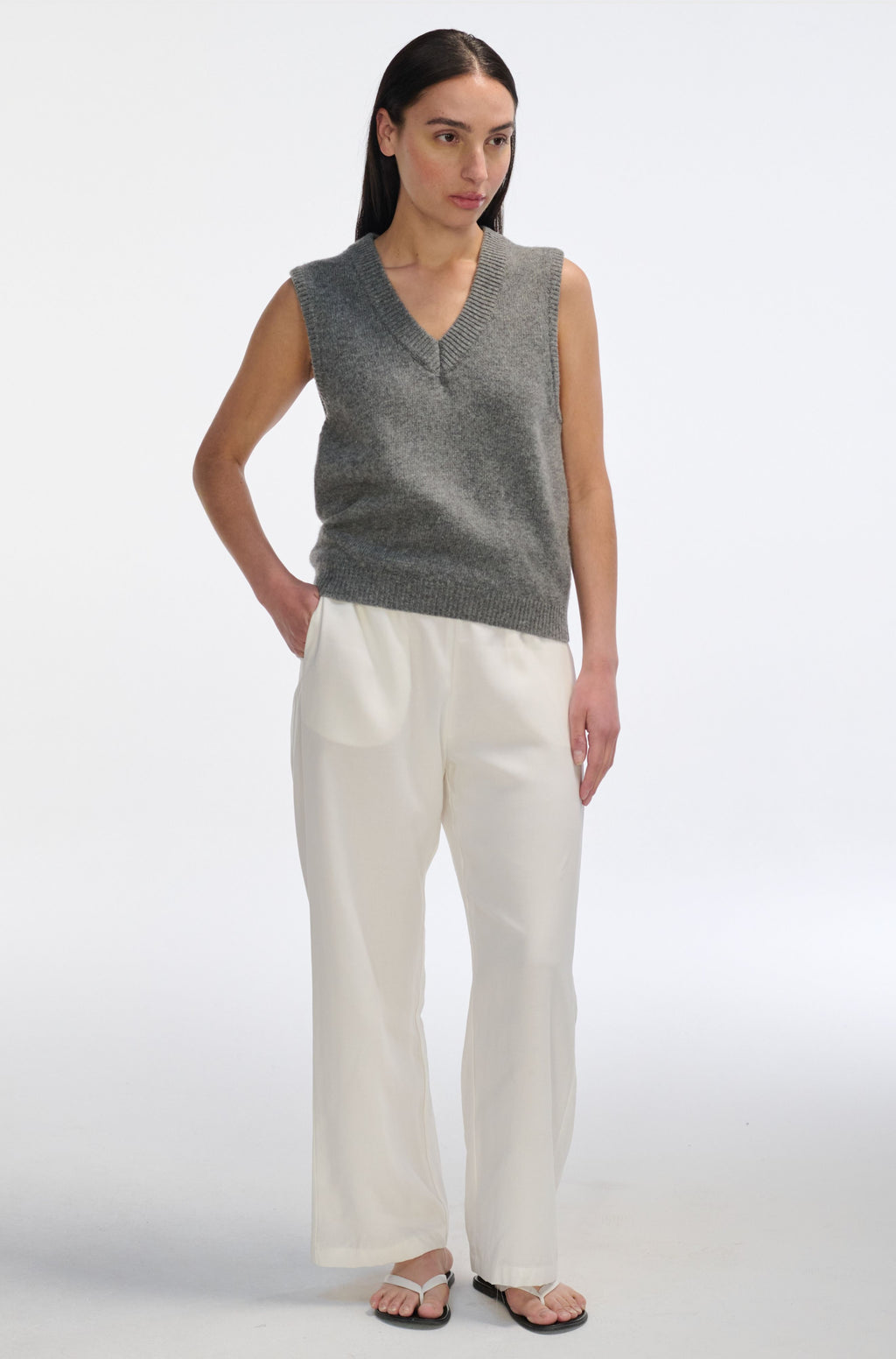 Woman wearing a gray sleeveless sweater and white pants on a white background