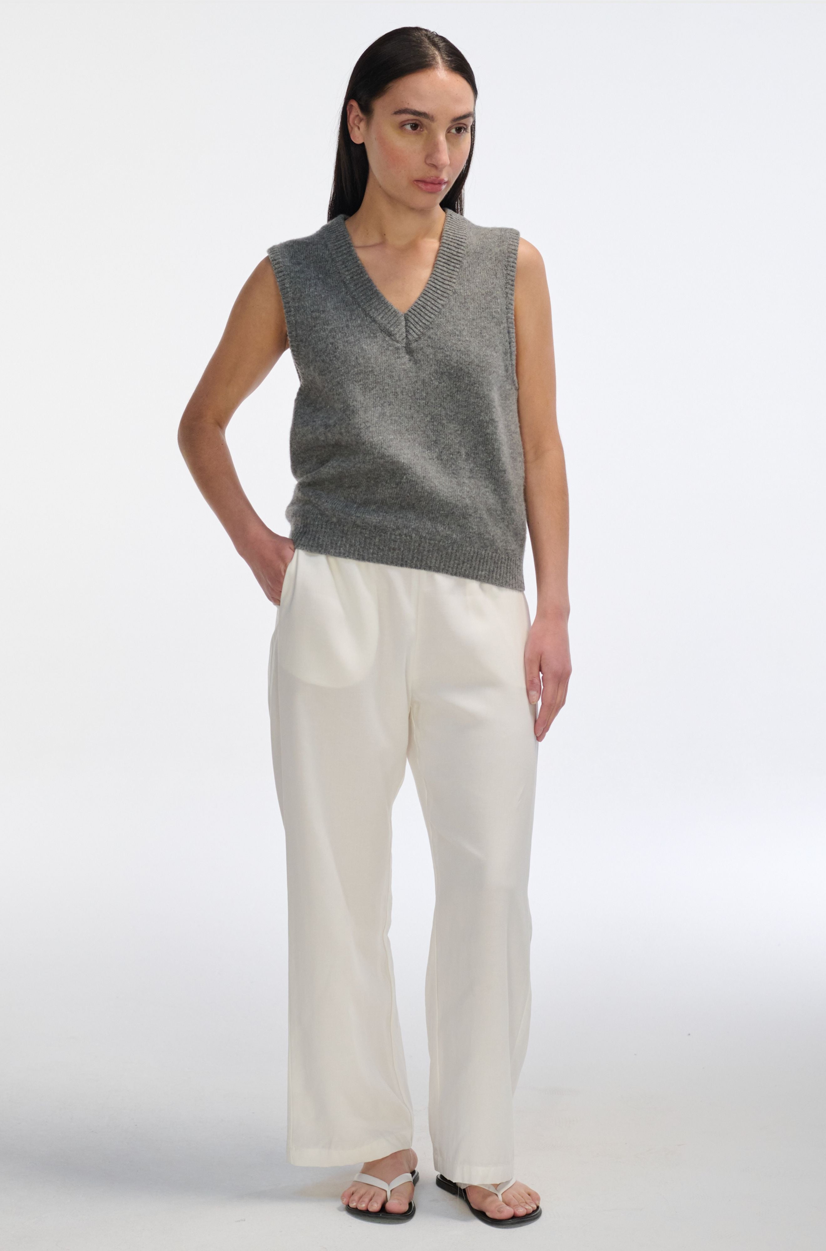 Woman wearing a gray sleeveless sweater and white pants on a white background