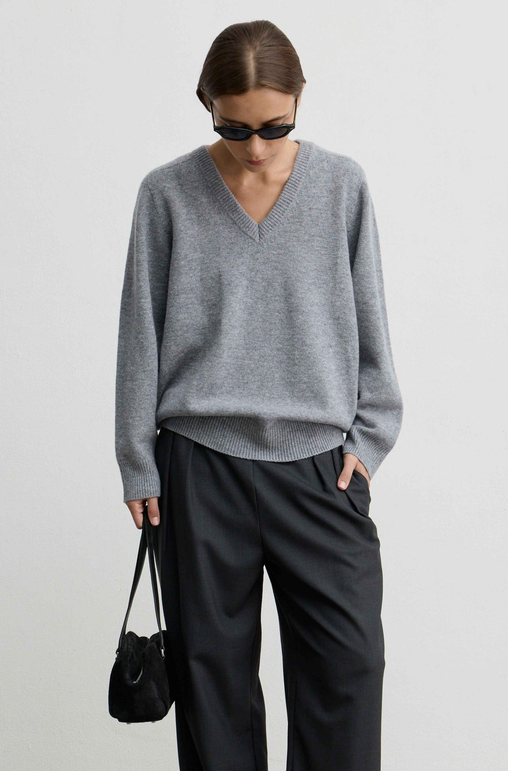 A model wearing a light gray v-neck knit sweater, black wide-leg trousers, black sunglasses, and carrying a small black shoulder bag, photographed against a plain light gray studio background.