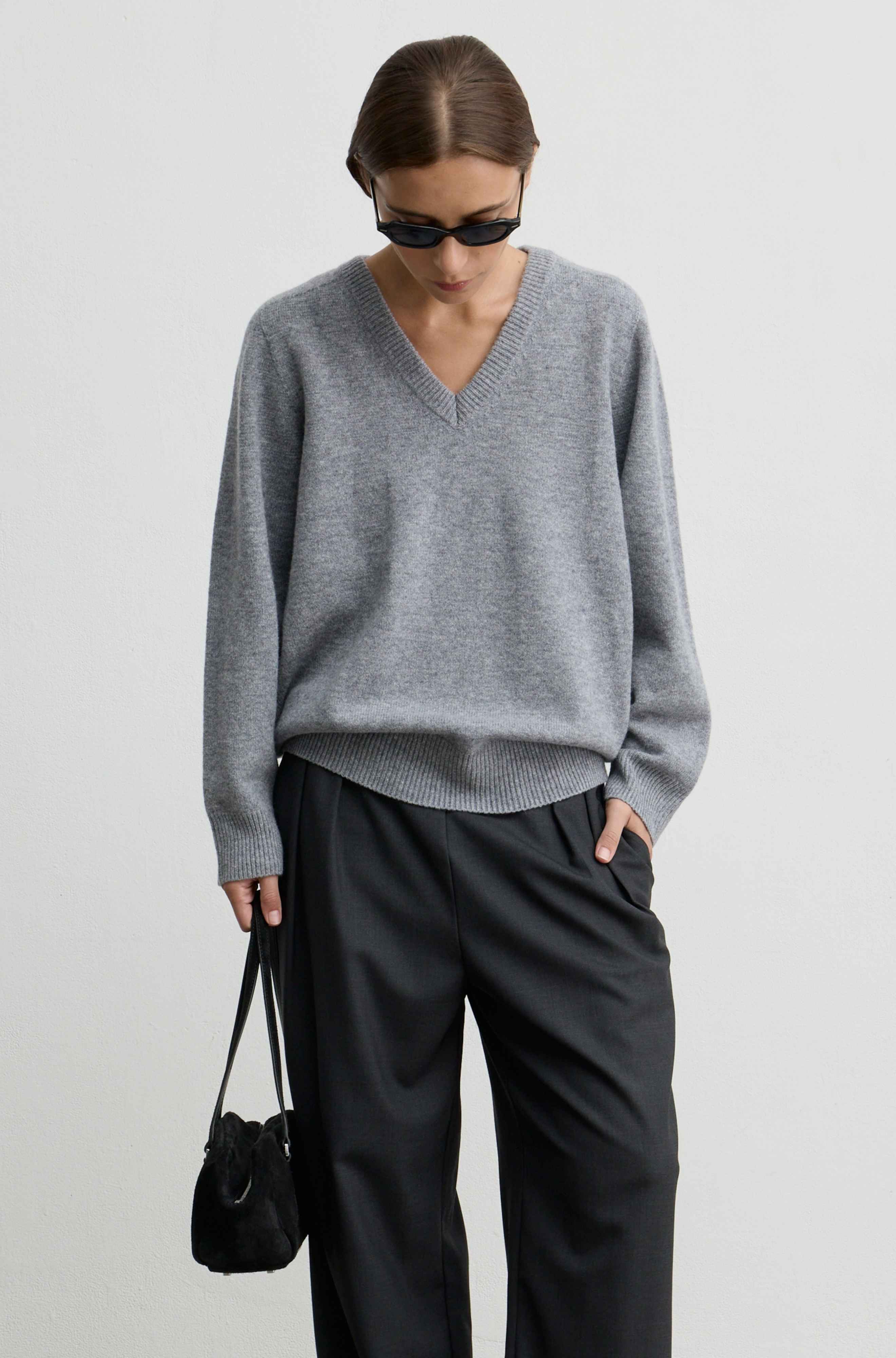 A model wearing a light gray v-neck knit sweater, black wide-leg trousers, black sunglasses, and carrying a small black shoulder bag, photographed against a plain light gray studio background.