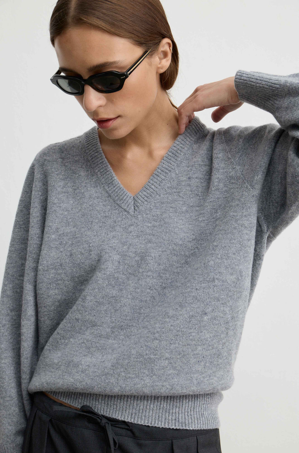 A woman wearing a light gray V-neck knit sweater and black sunglasses, shown from the chest up against a plain light background.