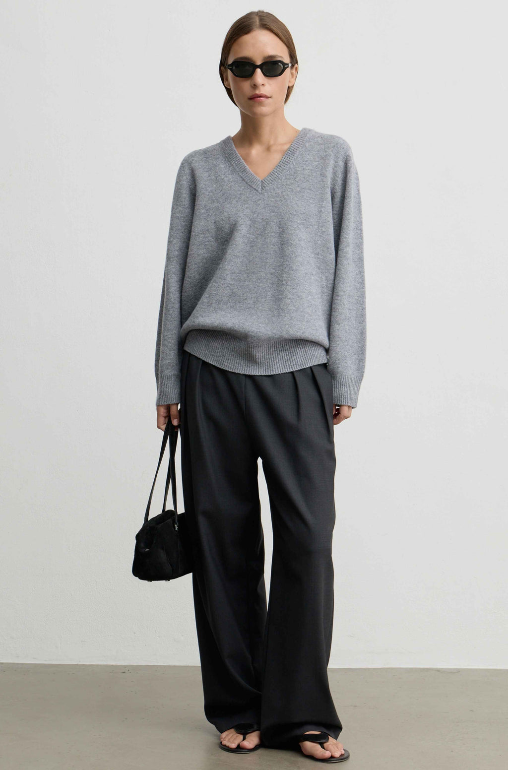 A model stands against a light gray studio wall wearing an oversized light gray V-neck sweater, loose black wide-leg pants, black sandals, and holding a small black handbag.