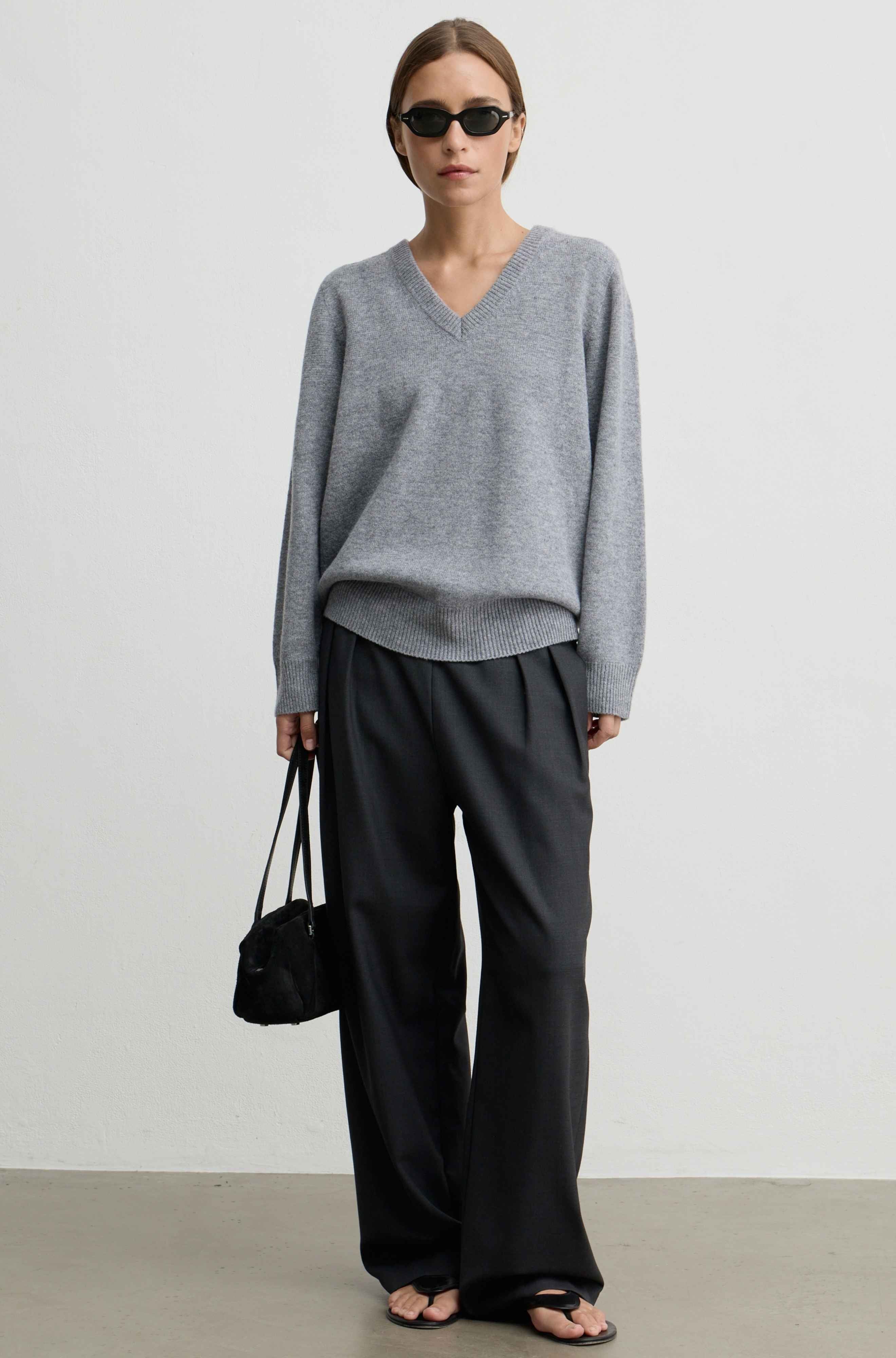 A model stands against a light gray studio wall wearing an oversized light gray V-neck sweater, loose black wide-leg pants, black sandals, and holding a small black handbag.