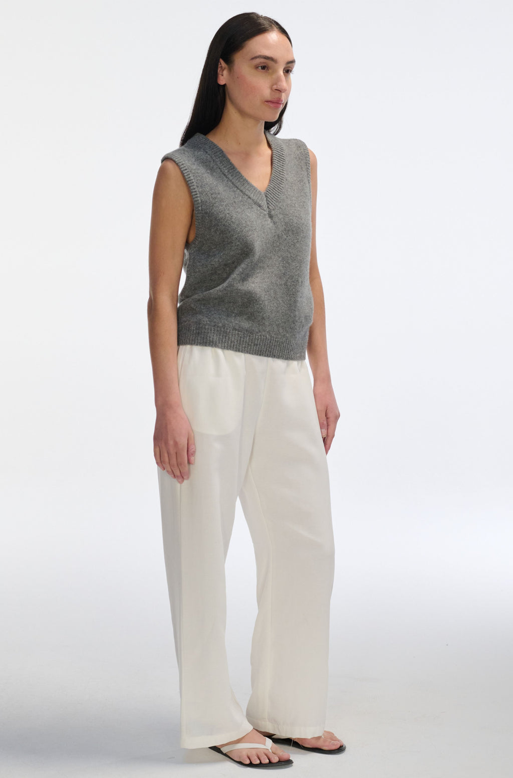Woman wearing a gray sleeveless sweater and white pants on a white background