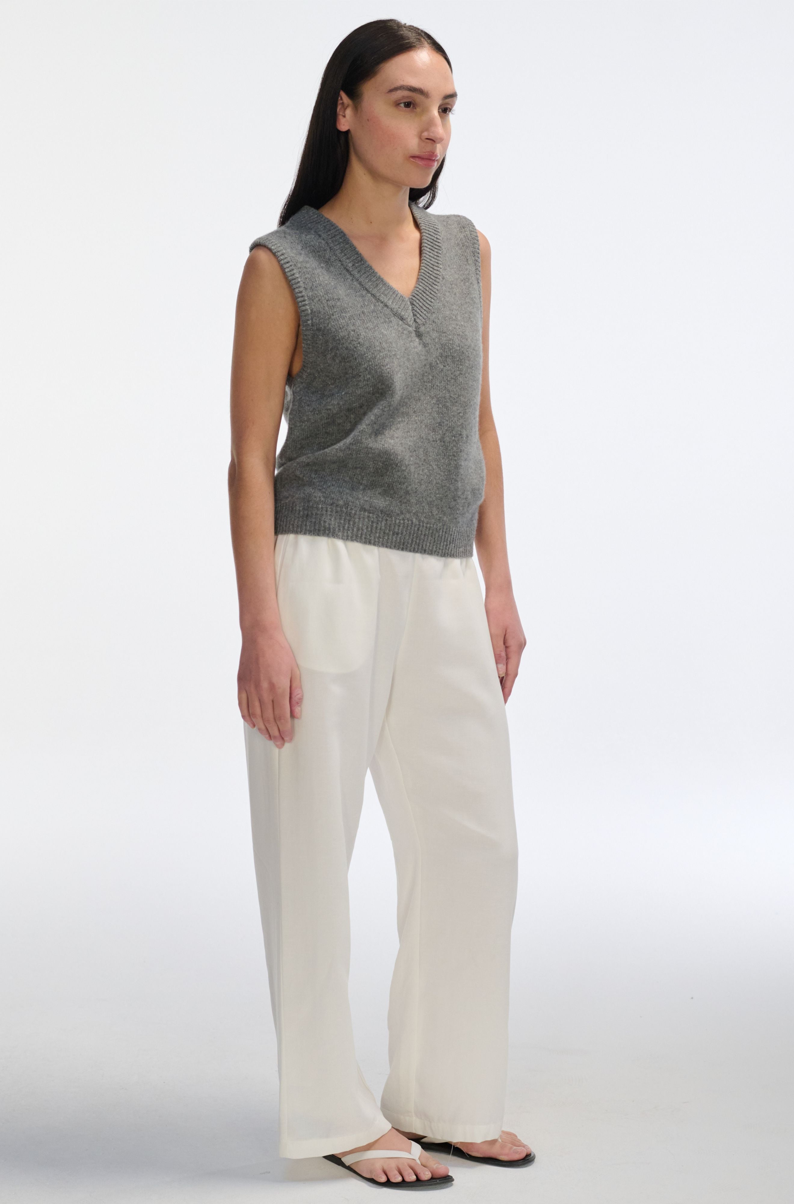 Woman wearing a gray sleeveless sweater and white pants on a white background