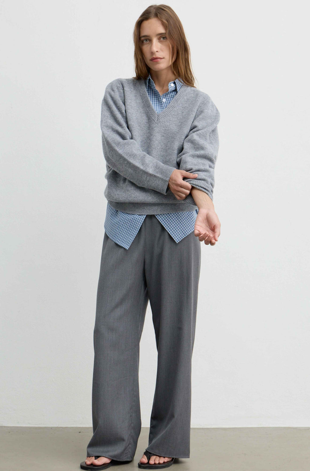 A woman modeling a gray knit V-neck sweater layered over a light blue and white checkered shirt, paired with wide-leg gray trousers and black sandals, against a plain white studio background.