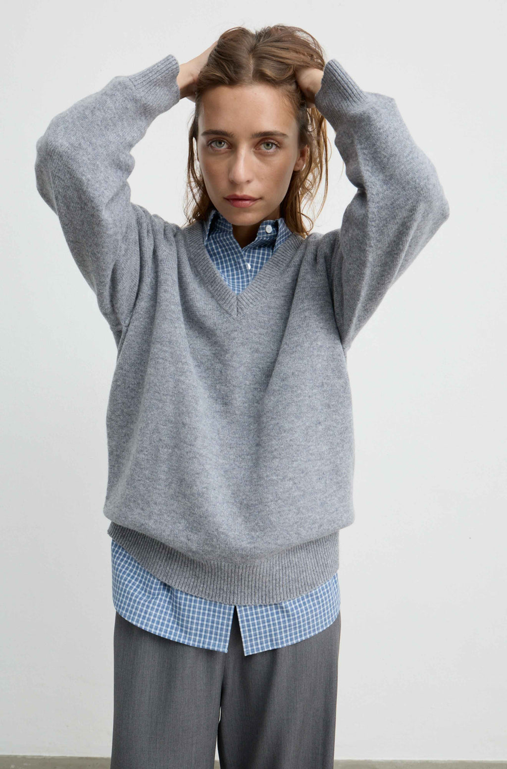A woman wearing a light gray v-neck knit sweater over a blue and white checkered shirt, paired with gray trousers, posing with her hands in her hair against a plain light gray background.