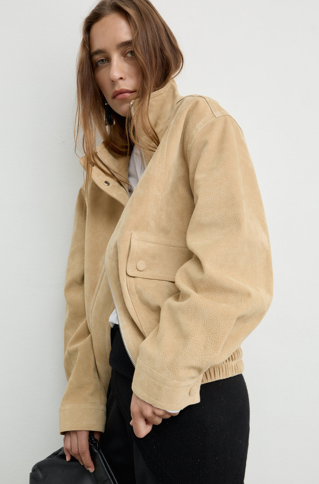 A woman wearing a beige suede jacket with a high collar, buttoned front, and flap pocket, paired with black pants, photographed against a plain light gray background.