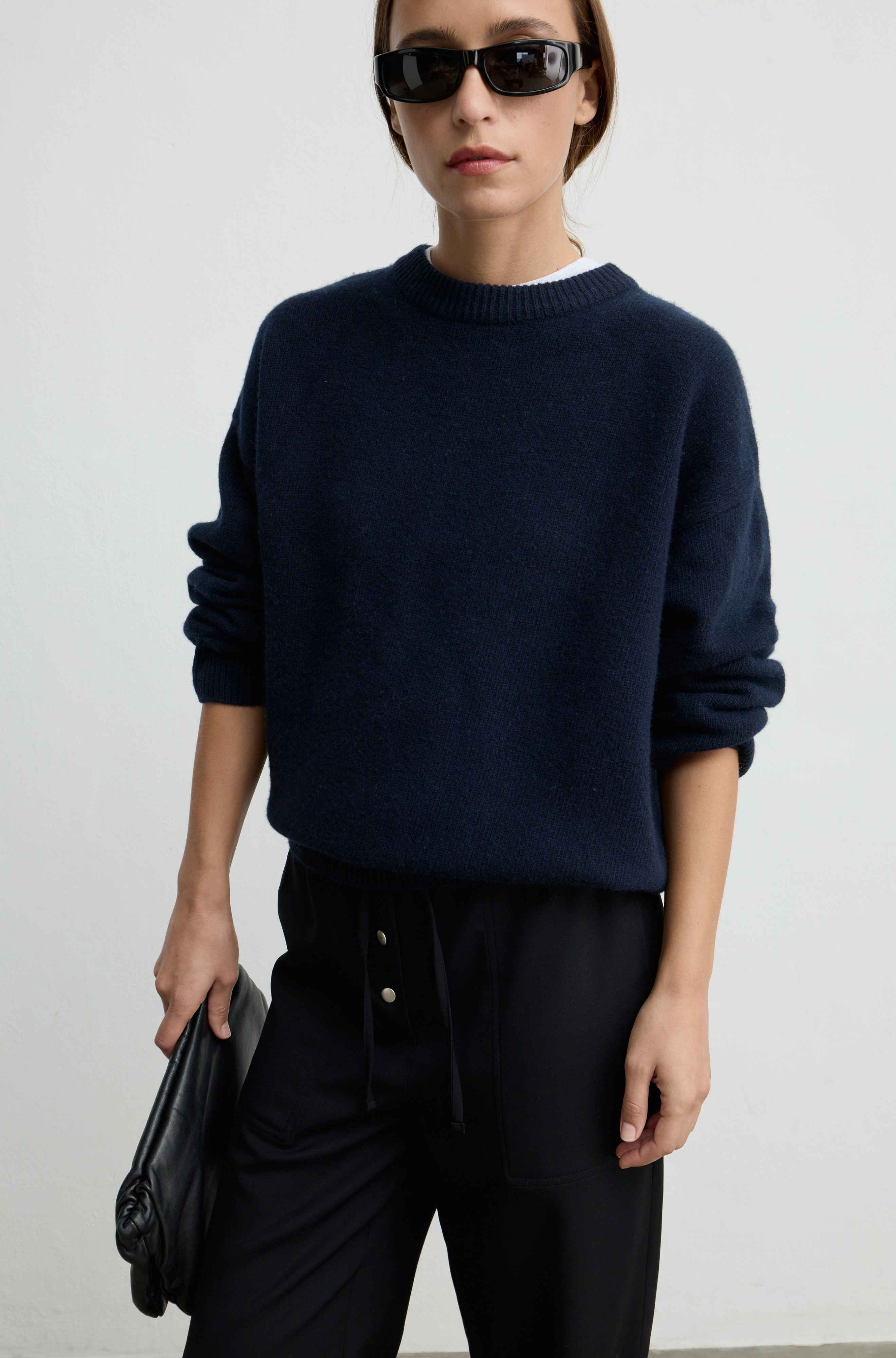 A woman wearing a dark navy blue knit crewneck sweater, black pants, and black sunglasses, holding a black clutch bag against a plain light gray background.