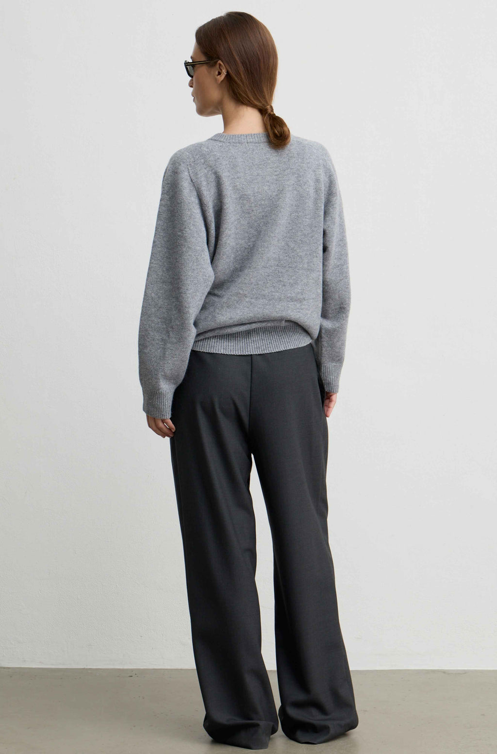 A woman shown from the back wearing a light gray knit sweater and dark charcoal wide-leg trousers, standing against a plain light gray wall.