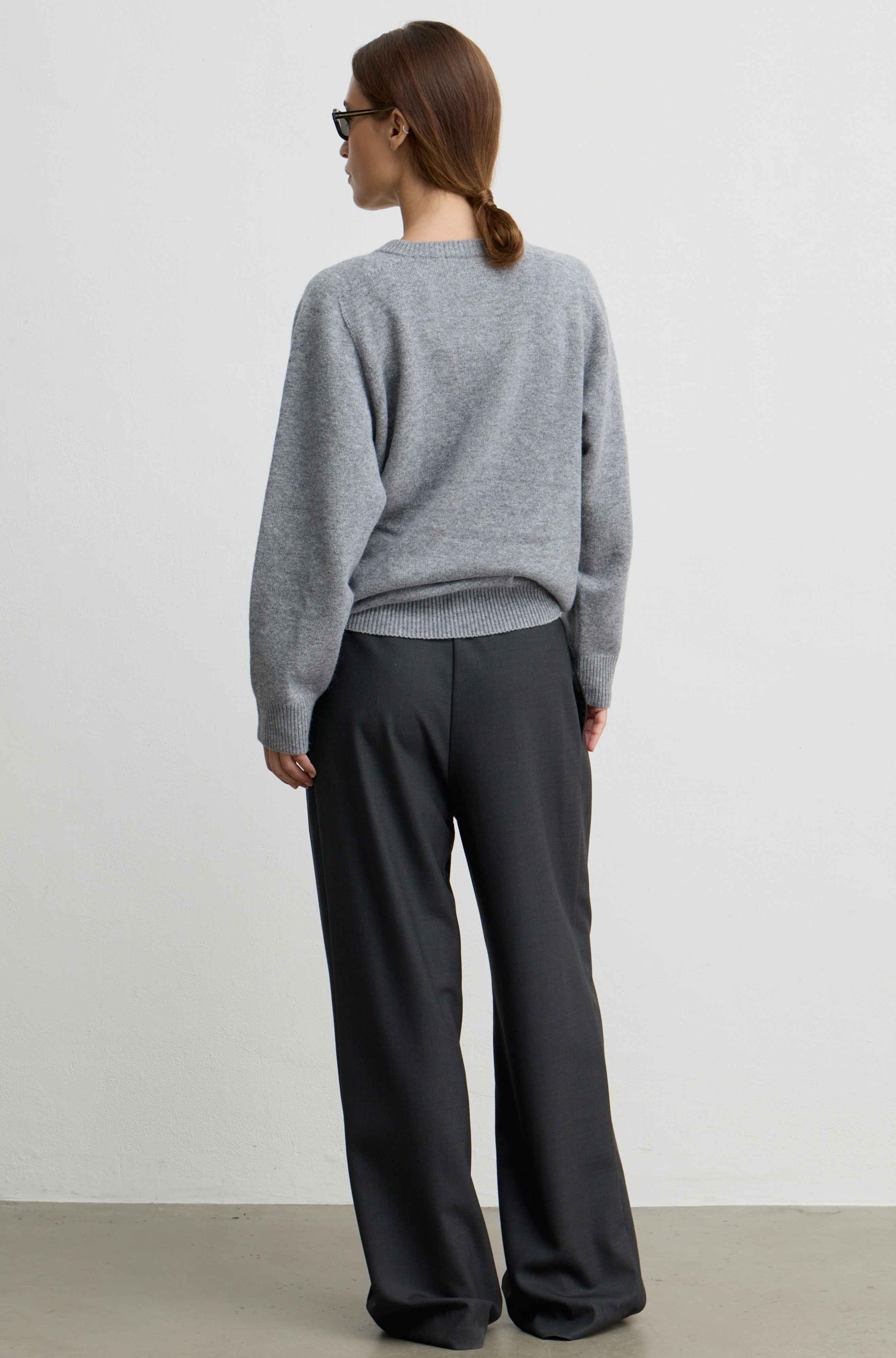 A woman shown from the back wearing a light gray knit sweater and dark charcoal wide-leg trousers, standing against a plain light gray wall.