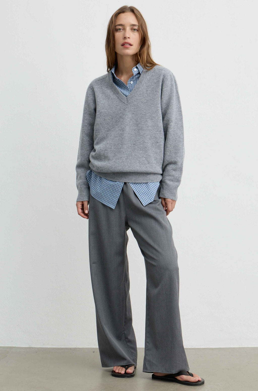 A woman stands against a plain light gray wall wearing a heather gray V-neck sweater over a blue-and-white gingham shirt, paired with loose gray trousers and black sandals.