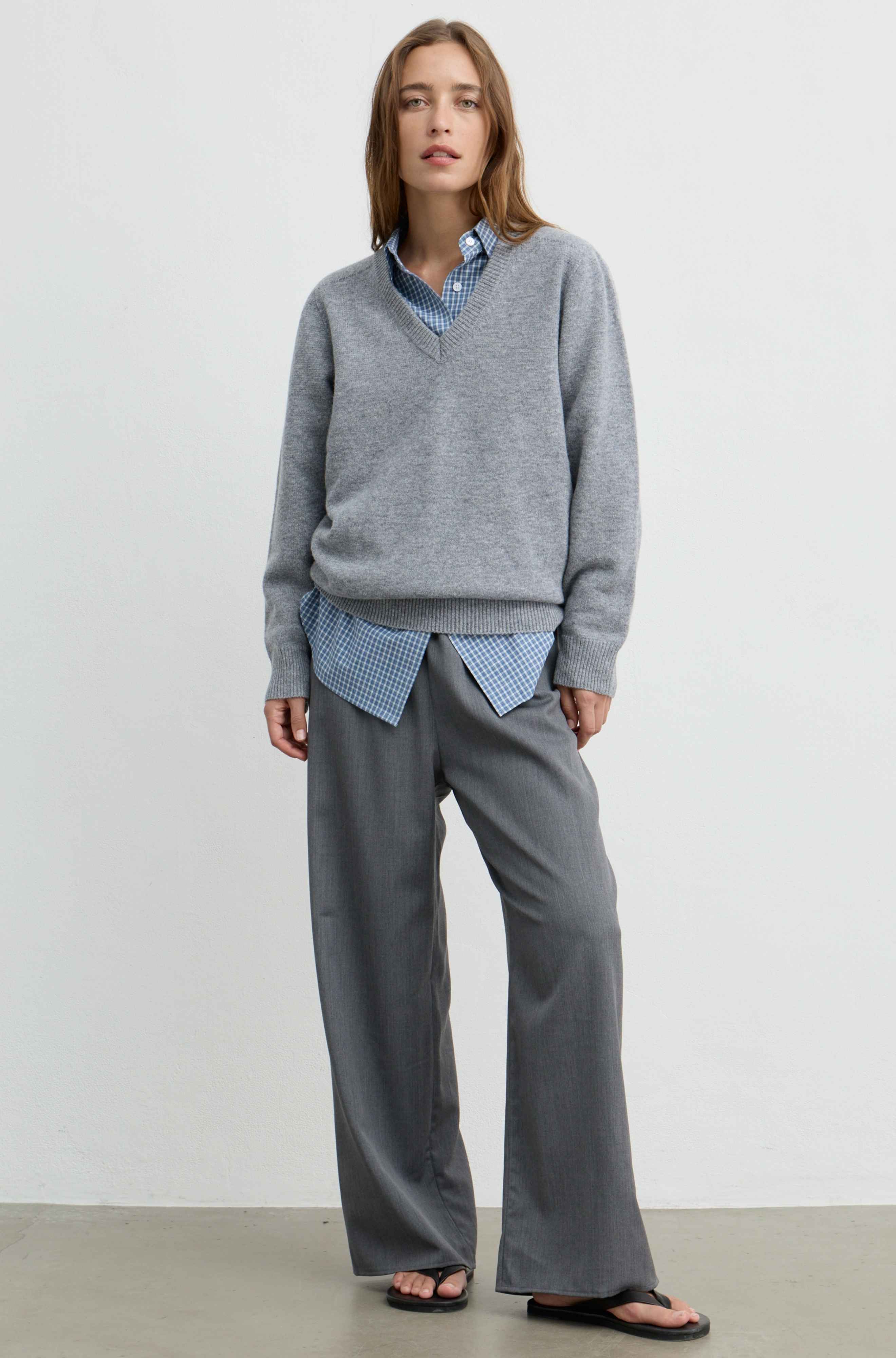 A woman stands against a plain light gray wall wearing a heather gray V-neck sweater over a blue-and-white gingham shirt, paired with loose gray trousers and black sandals.