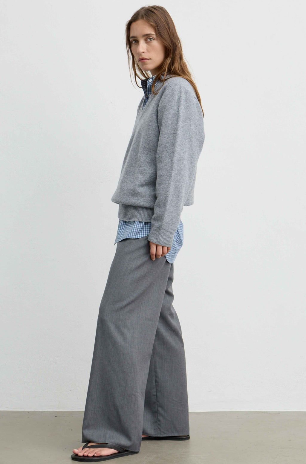 A woman modeling a gray knit crewneck sweater layered over a blue and white gingham shirt, paired with wide-leg gray trousers and black flip-flops, standing against a plain light gray studio background.
