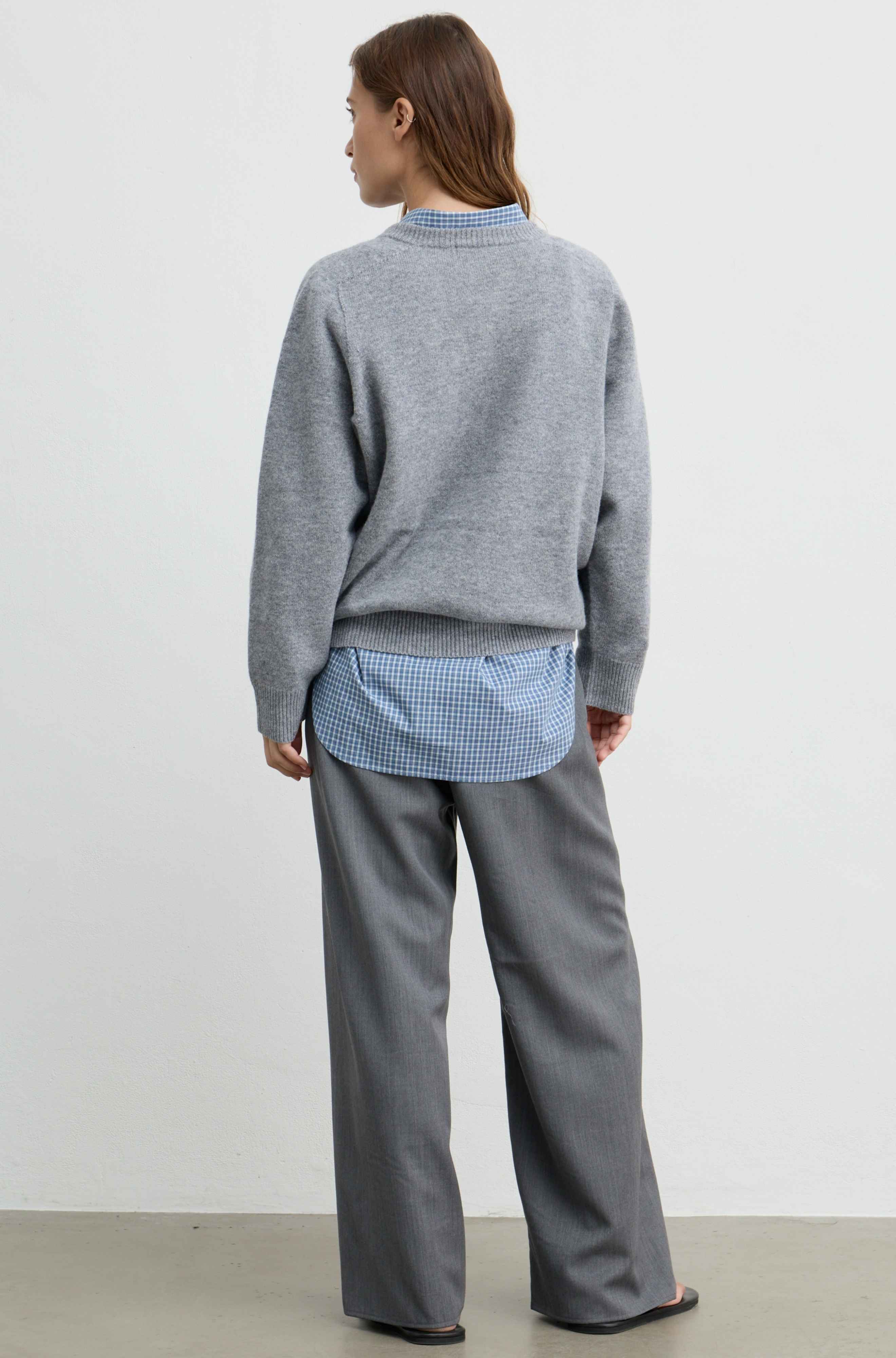A woman is shown from the back wearing a heather gray crewneck sweater layered over a light blue and white gingham shirt, paired with wide-leg gray trousers, against a plain white studio background.