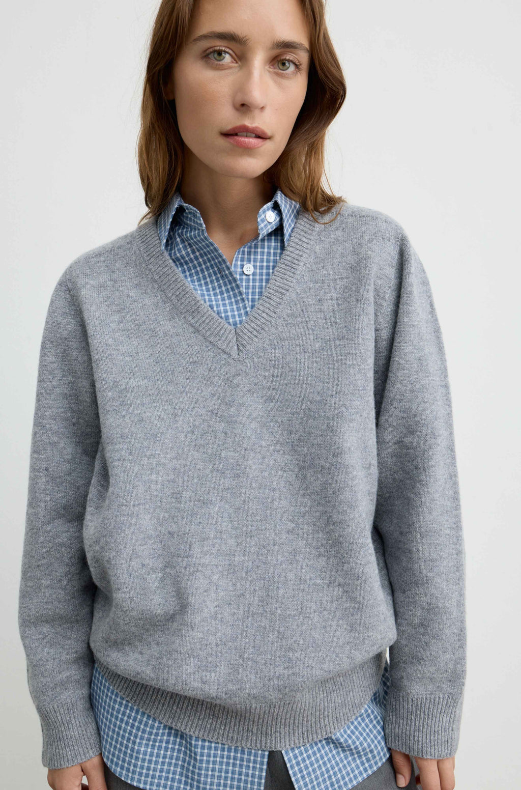 A woman wearing a light gray v-neck knit sweater over a blue and white checkered button-down shirt, photographed against a plain light gray studio background.