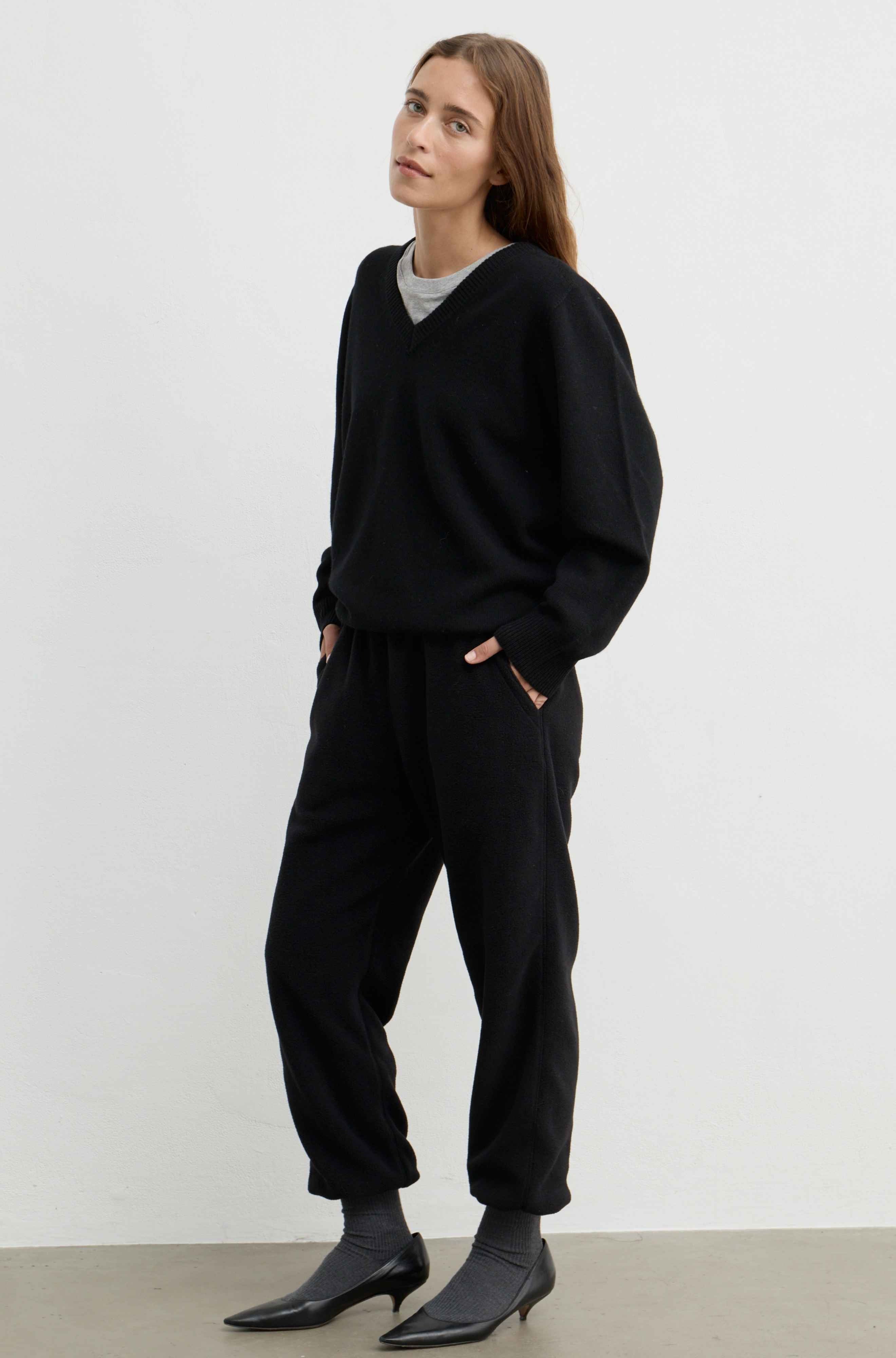A woman modeling a black knit lounge set consisting of a V-neck sweater and matching jogger pants, styled over a light gray t-shirt and black pointed-toe ankle boots against a plain white studio background.