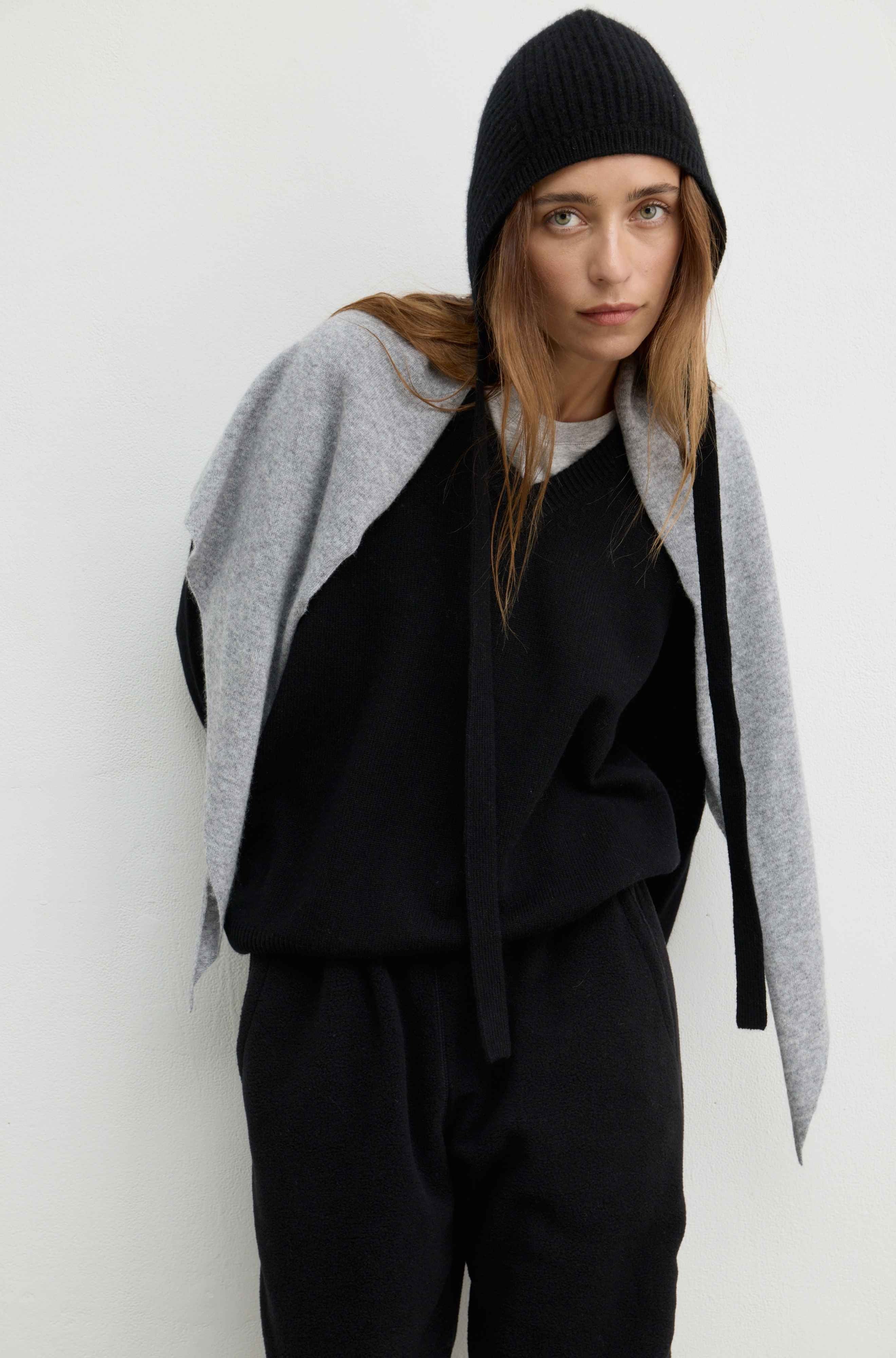 A model wearing a black knit beanie, a black sweatshirt with a gray cape-style sleeve overlay, and black sweatpants, posed against a plain off-white background.