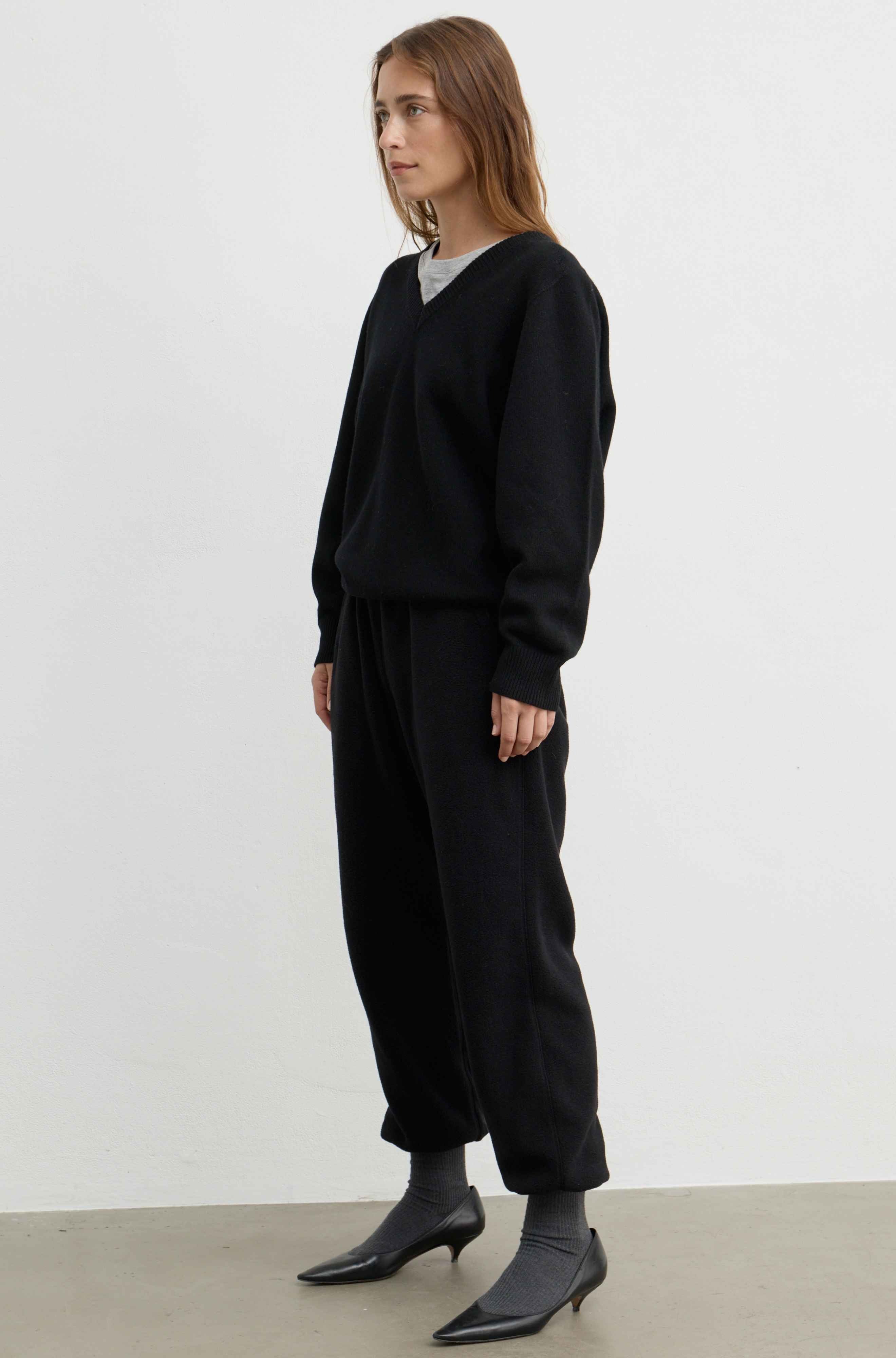 A woman modeling a black knit sweater and matching black wide-leg pants, paired with black pointed-toe ankle boots, against a plain light gray studio background.