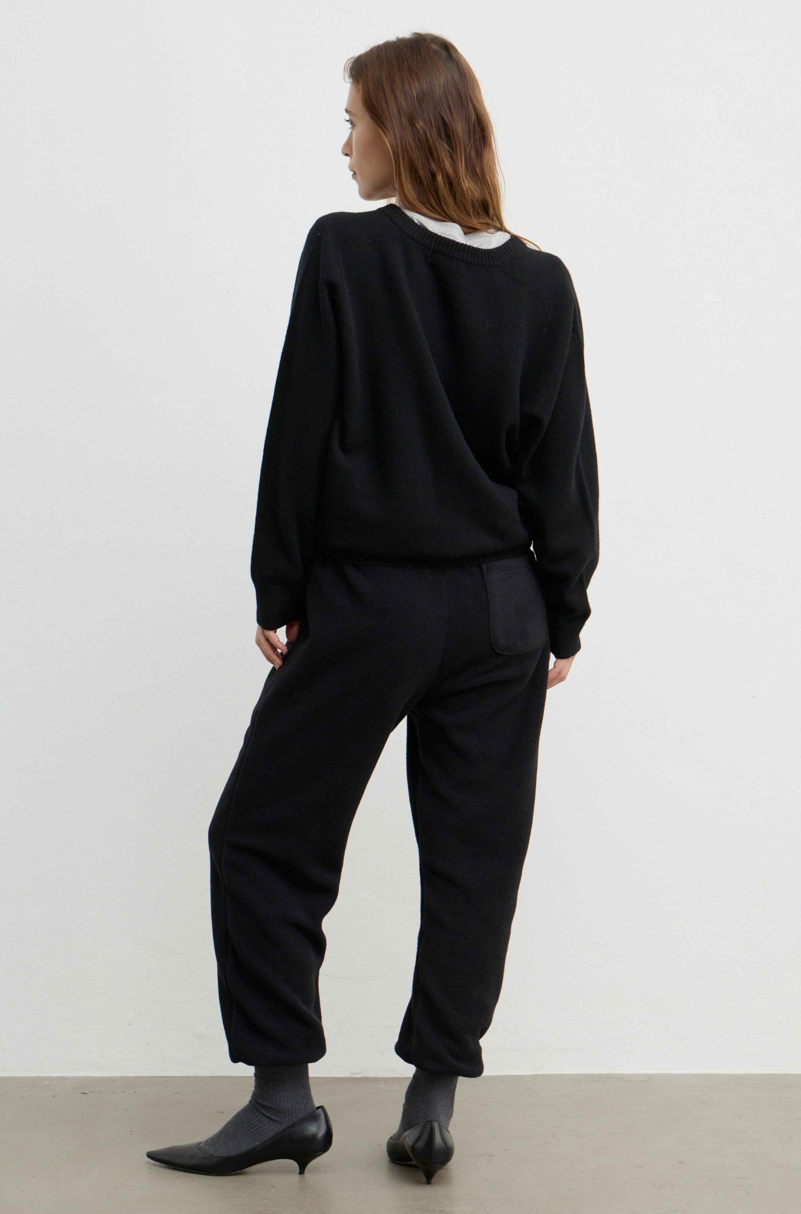 A woman is shown from behind wearing a black crewneck sweatshirt, black wide-leg cropped pants, black socks, and black pointed-toe heels against a plain white studio background.