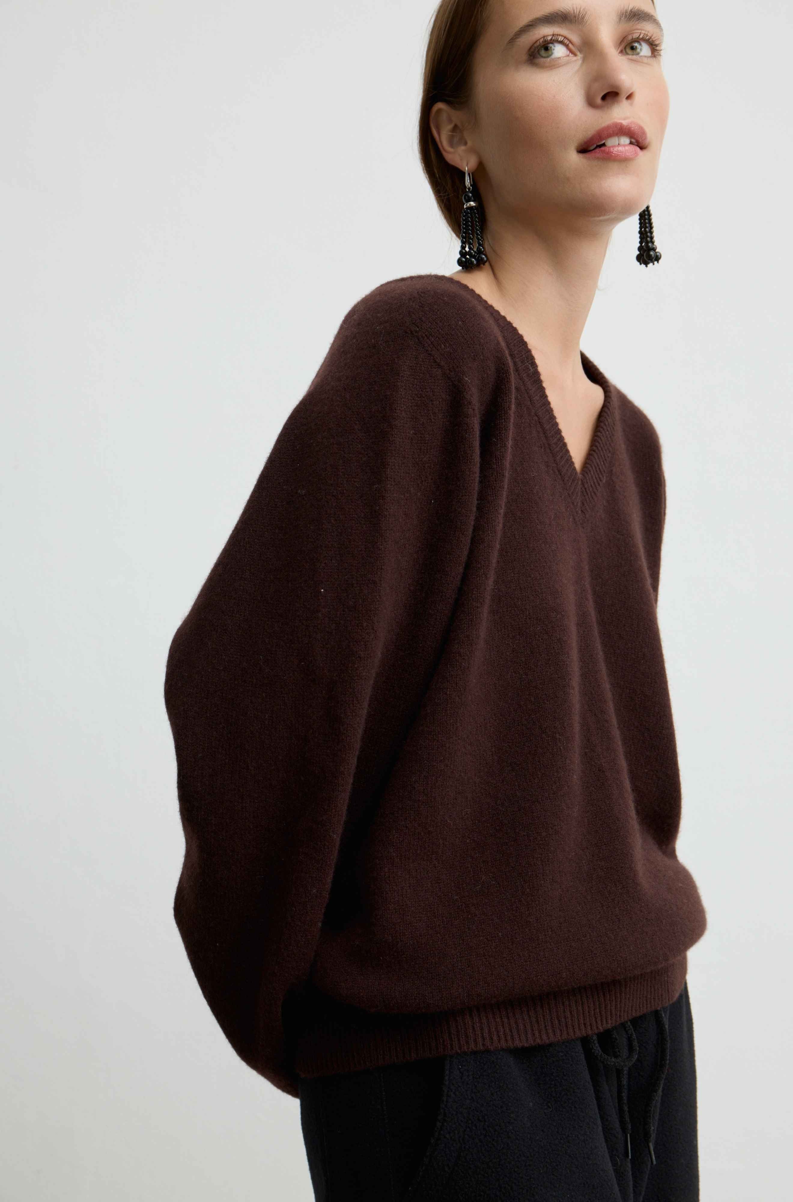 A woman wearing a dark brown knit sweater with a wide V-neck and oversized sleeves, paired with dark pants, photographed against a plain light gray background.