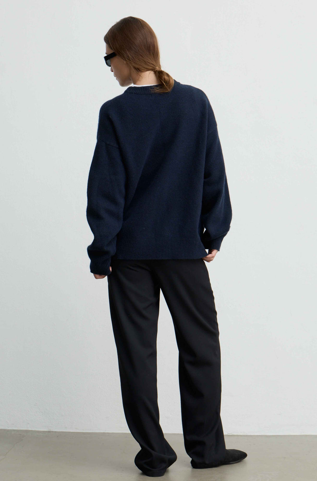 A person shown from the back wearing an oversized dark navy knit sweater and loose black trousers, standing against a plain light gray wall.