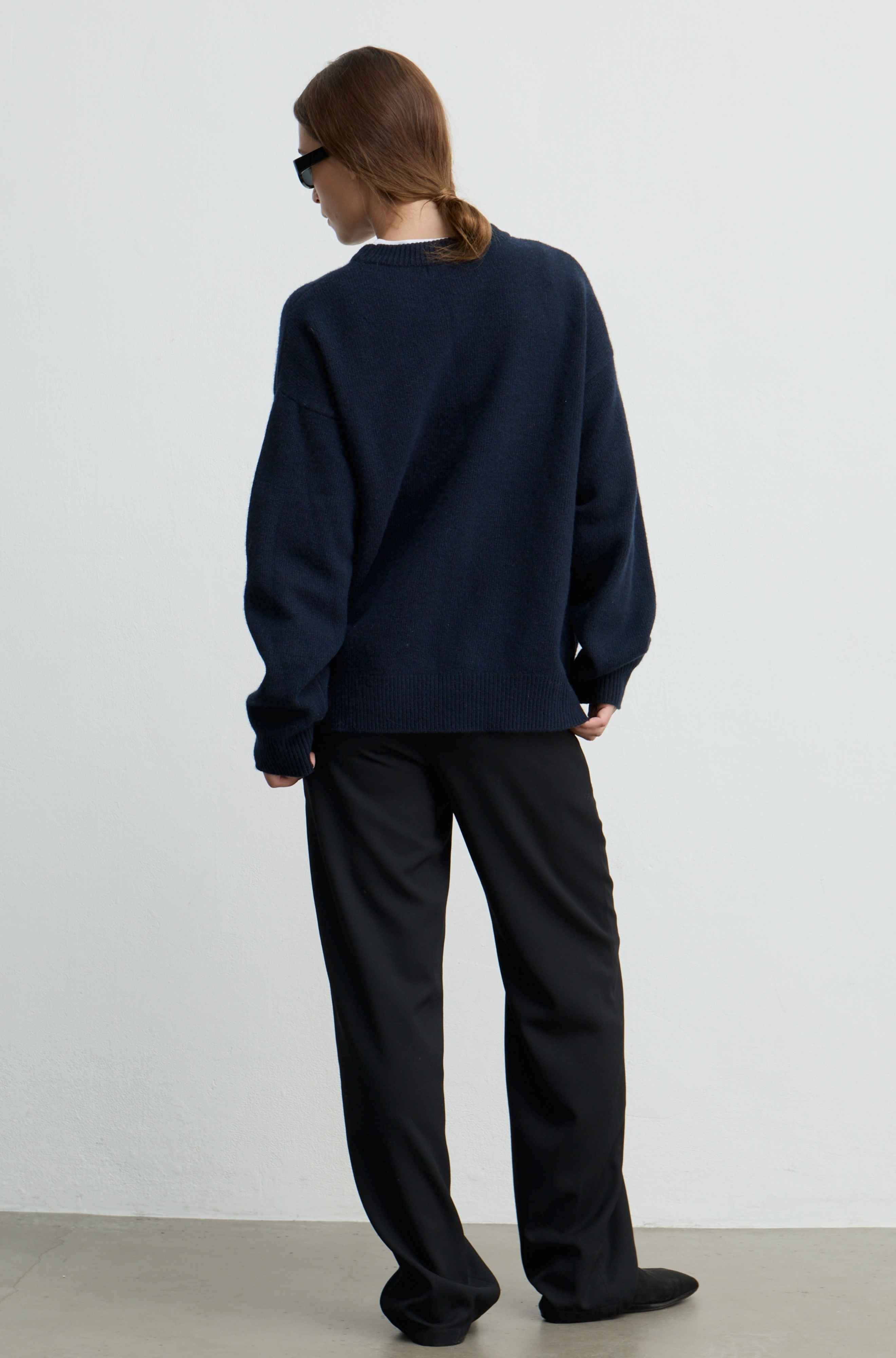 A person shown from the back wearing an oversized dark navy knit sweater and loose black trousers, standing against a plain light gray wall.