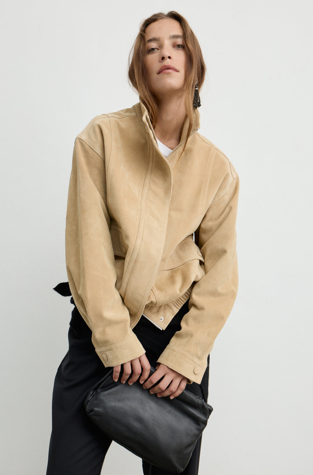 A woman wearing a beige suede jacket with a high collar and asymmetrical front closure, paired with black pants and holding a black leather clutch bag, set against a plain light gray studio background.