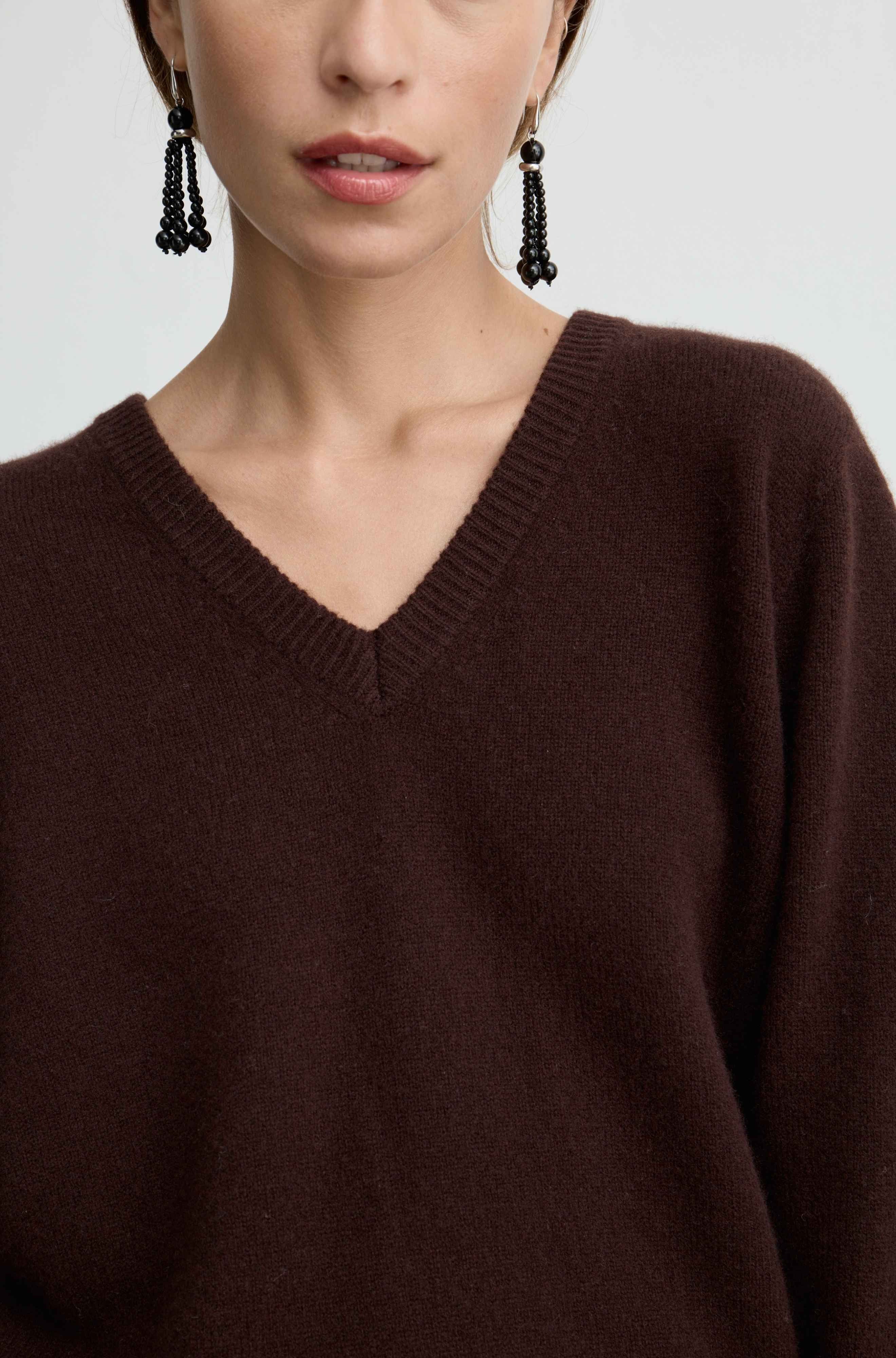 A close-up of a model wearing a dark brown V-neck knit sweater and black beaded drop earrings against a light gray background.