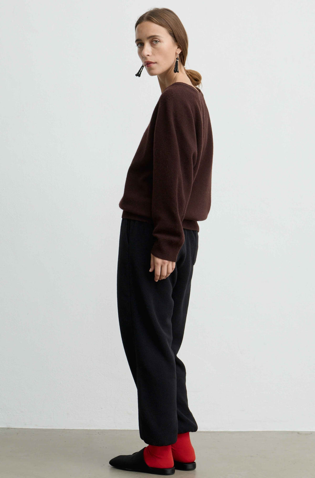 A woman stands in profile wearing a dark brown knit sweater, loose black trousers, bright red socks, and black slip-on shoes against a plain light gray studio background.