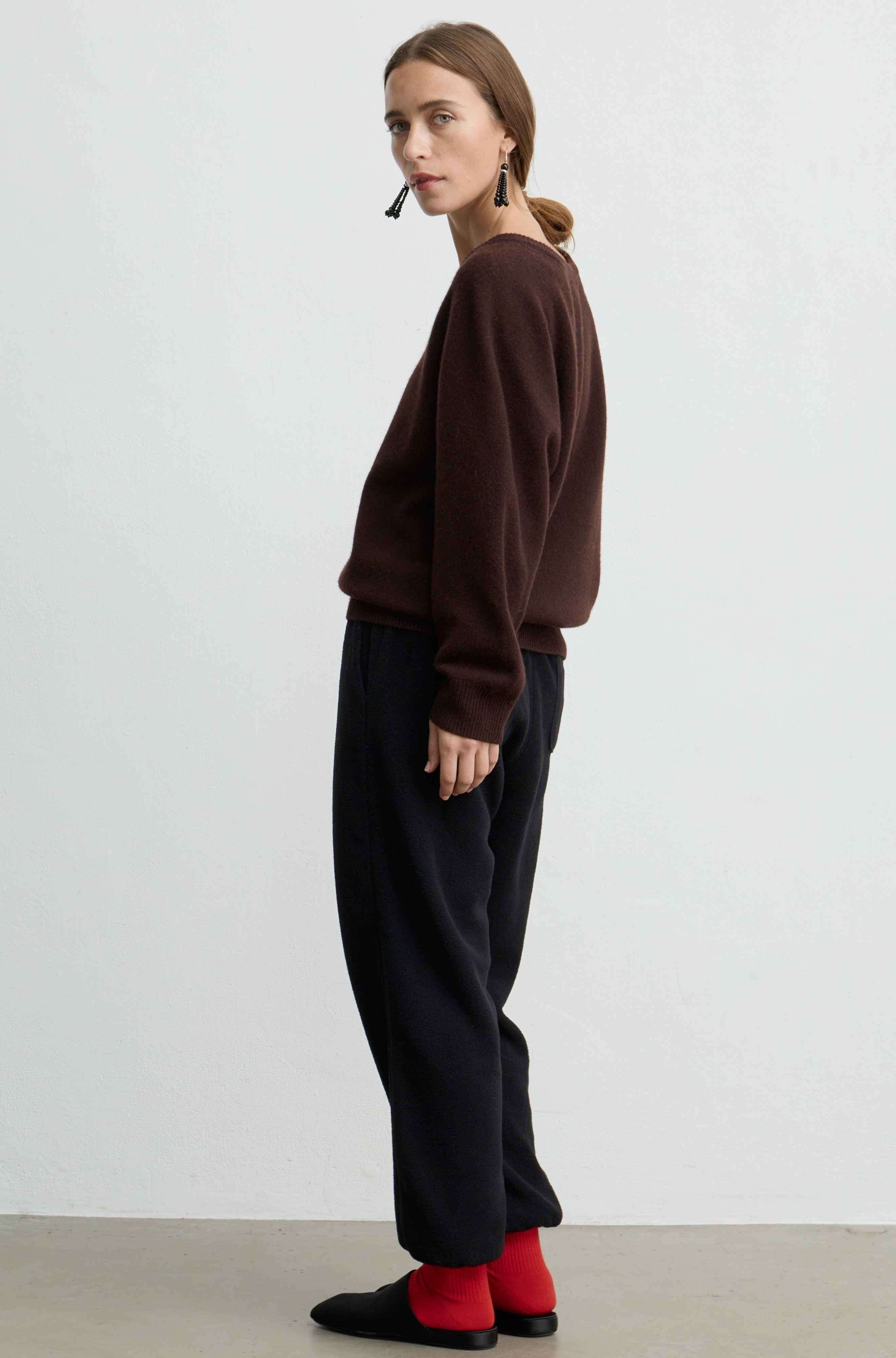 A woman stands in profile wearing a dark brown knit sweater, loose black trousers, bright red socks, and black slip-on shoes against a plain light gray studio background.