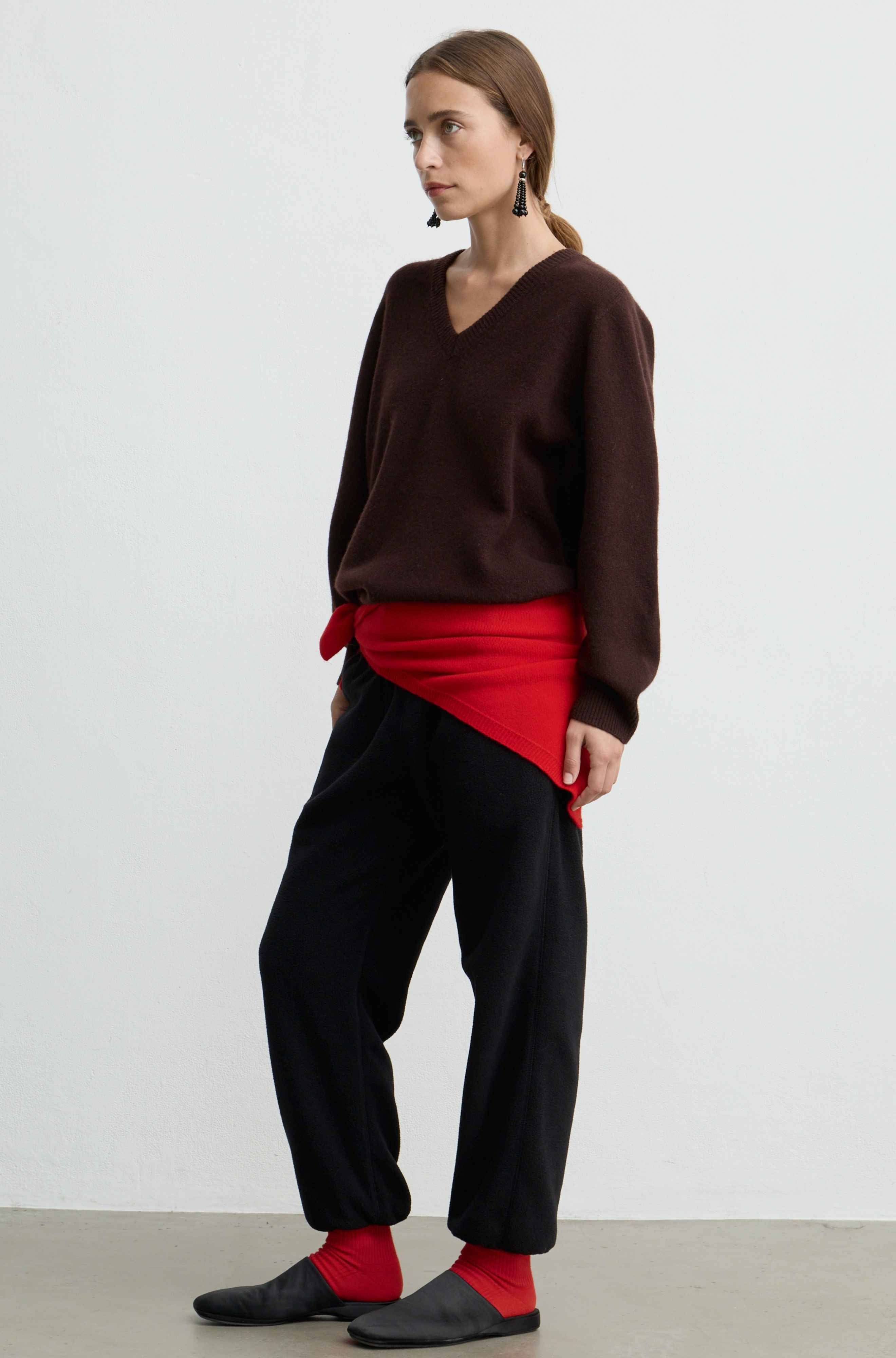 A woman stands in profile wearing a dark brown V-neck sweater, black wide-leg pants, and red socks, with a bright red fabric wrapped around her waist. She is photographed against a plain light gray studio background.